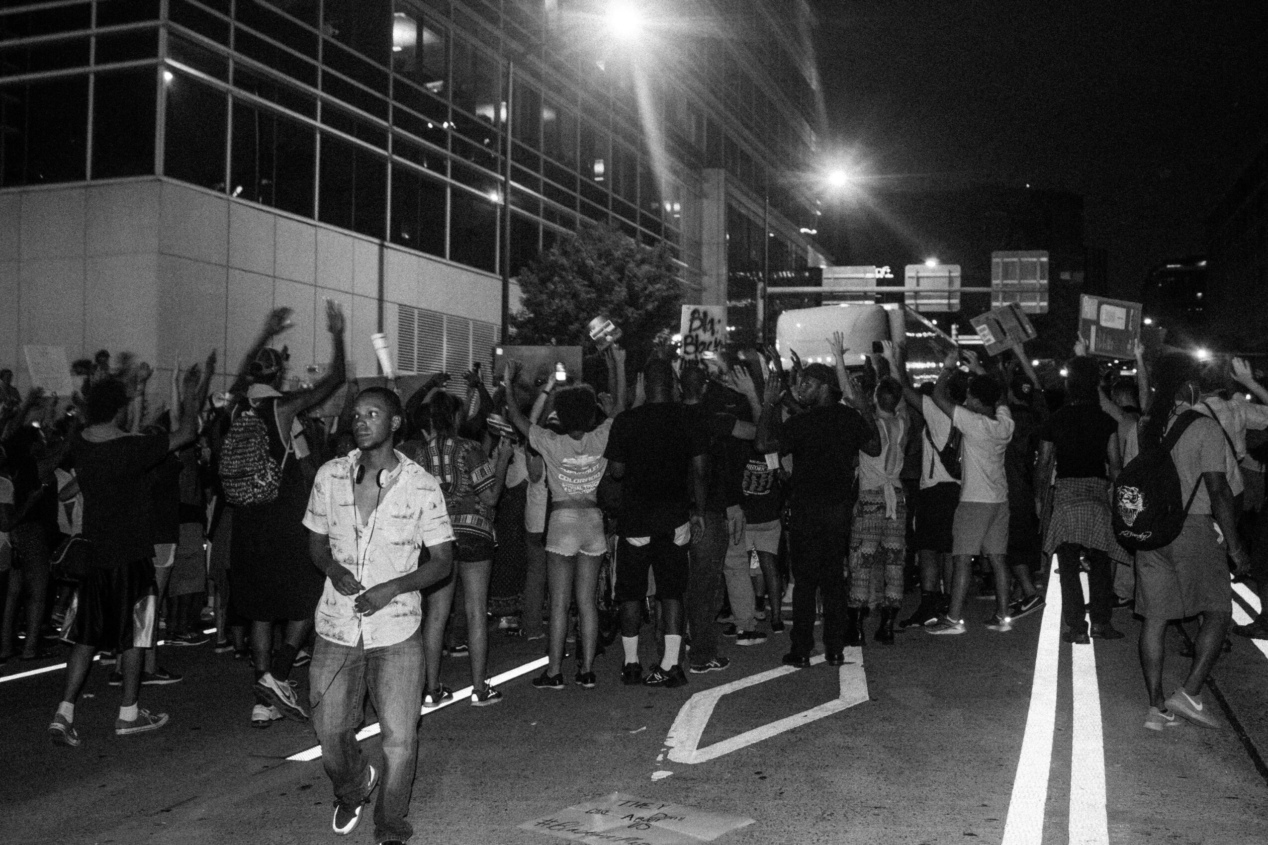Crowd of people gathered outside a building at night, some raising their hands, with streetlights and vehicles in the background.