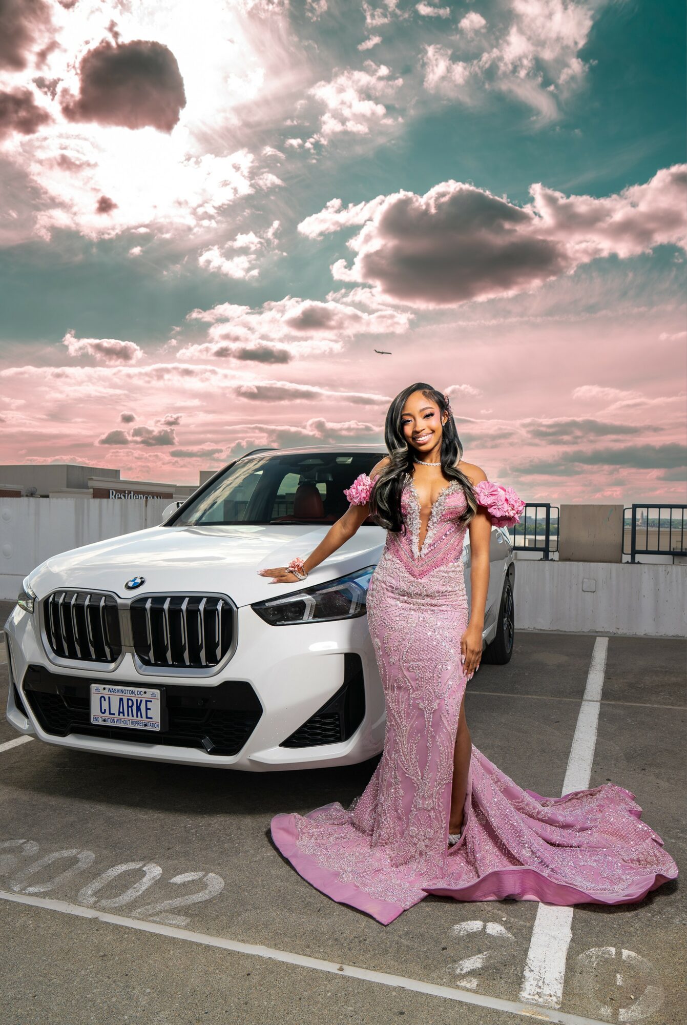 Woman in pink gown standing next to a white car on rooftop parking lot, sky with clouds in background.