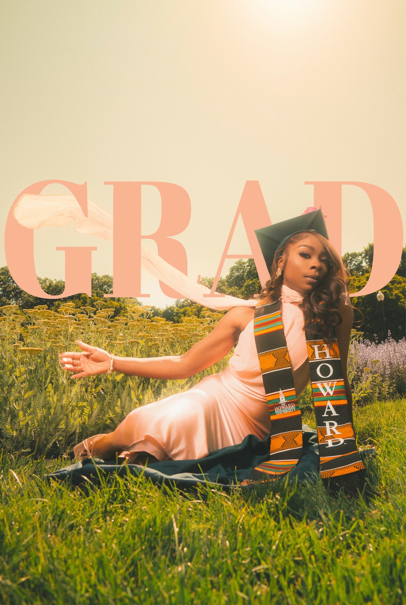 Graduate in cap and gown sitting on grass in a field, with large pink 'GRAD' text overlay.