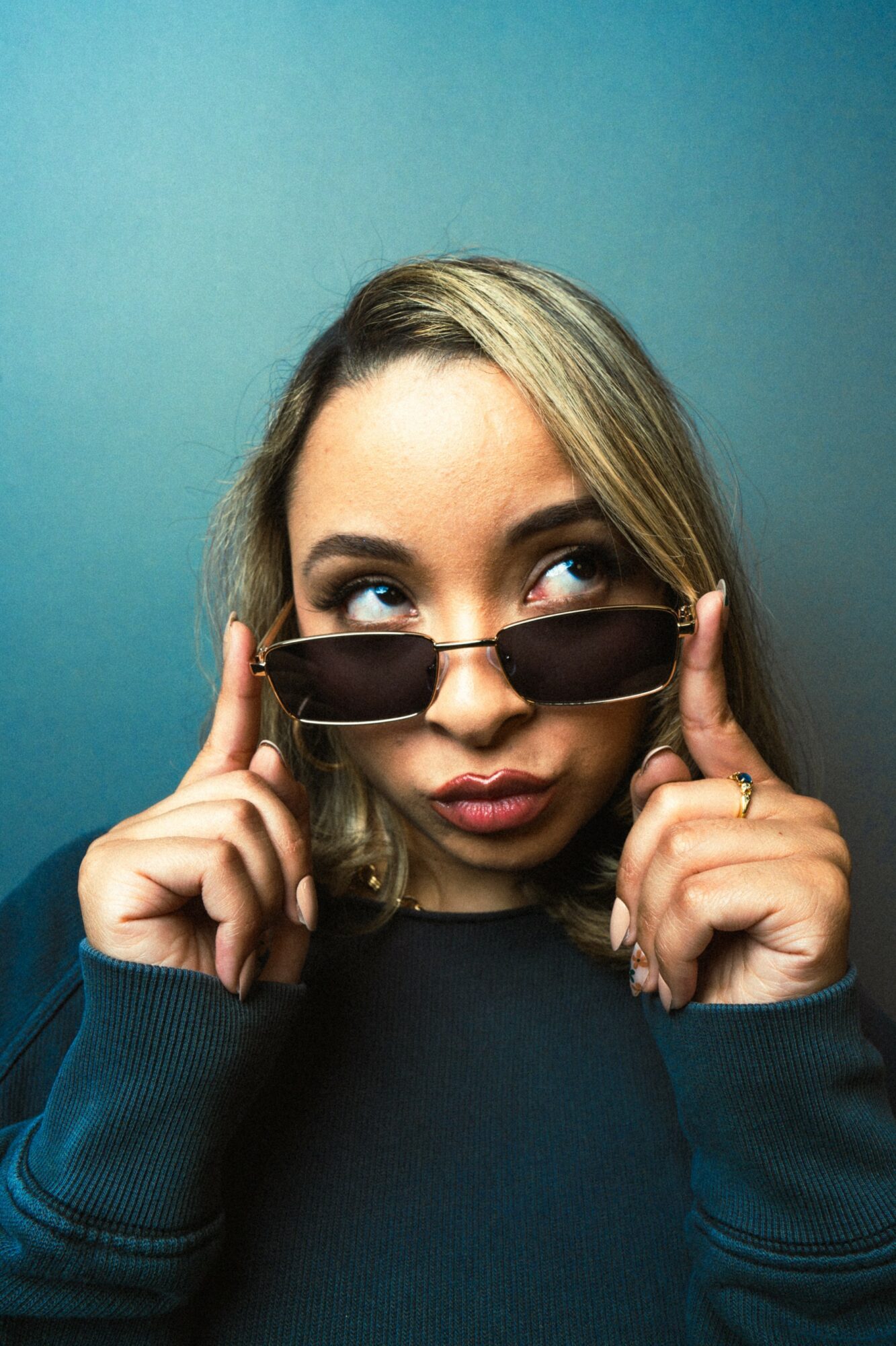 Young woman with blonde hair wearing sunglasses, looking over them, against a blue background.