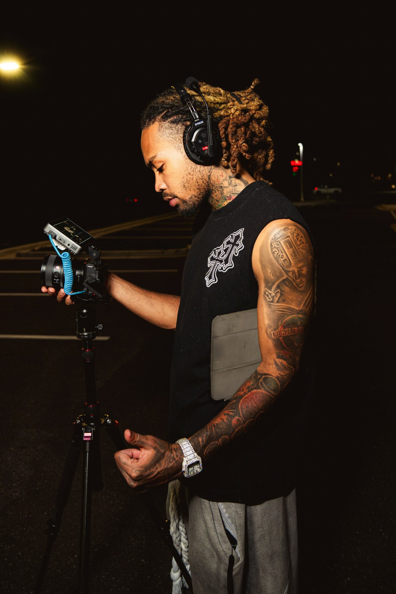 Young man with dreadlocks and tattoos wearing headphones, holding a device, standing outdoors at night.