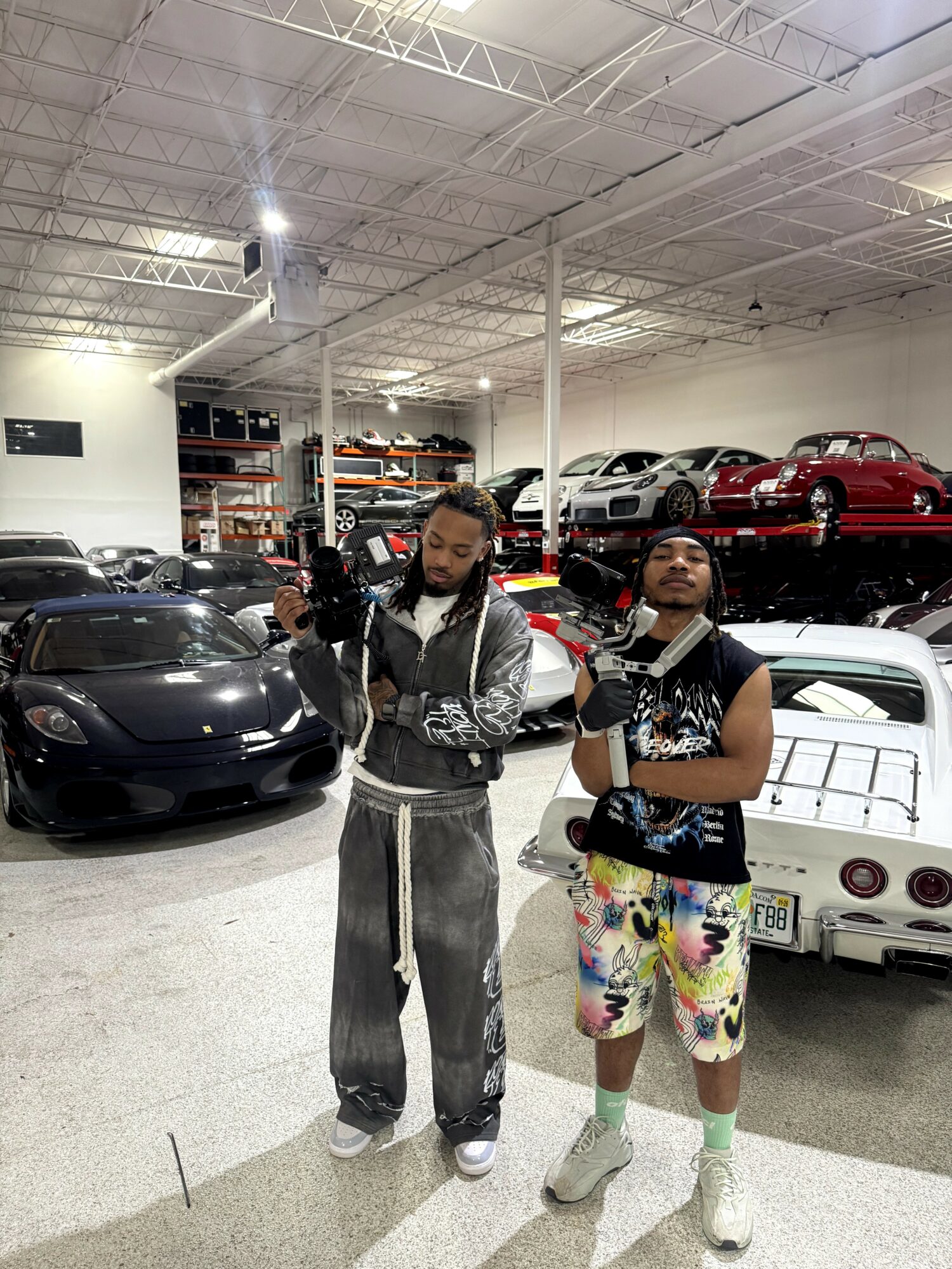 Two young men stand in a car showroom with sports cars and classic cars in the background, holding cameras and bags.