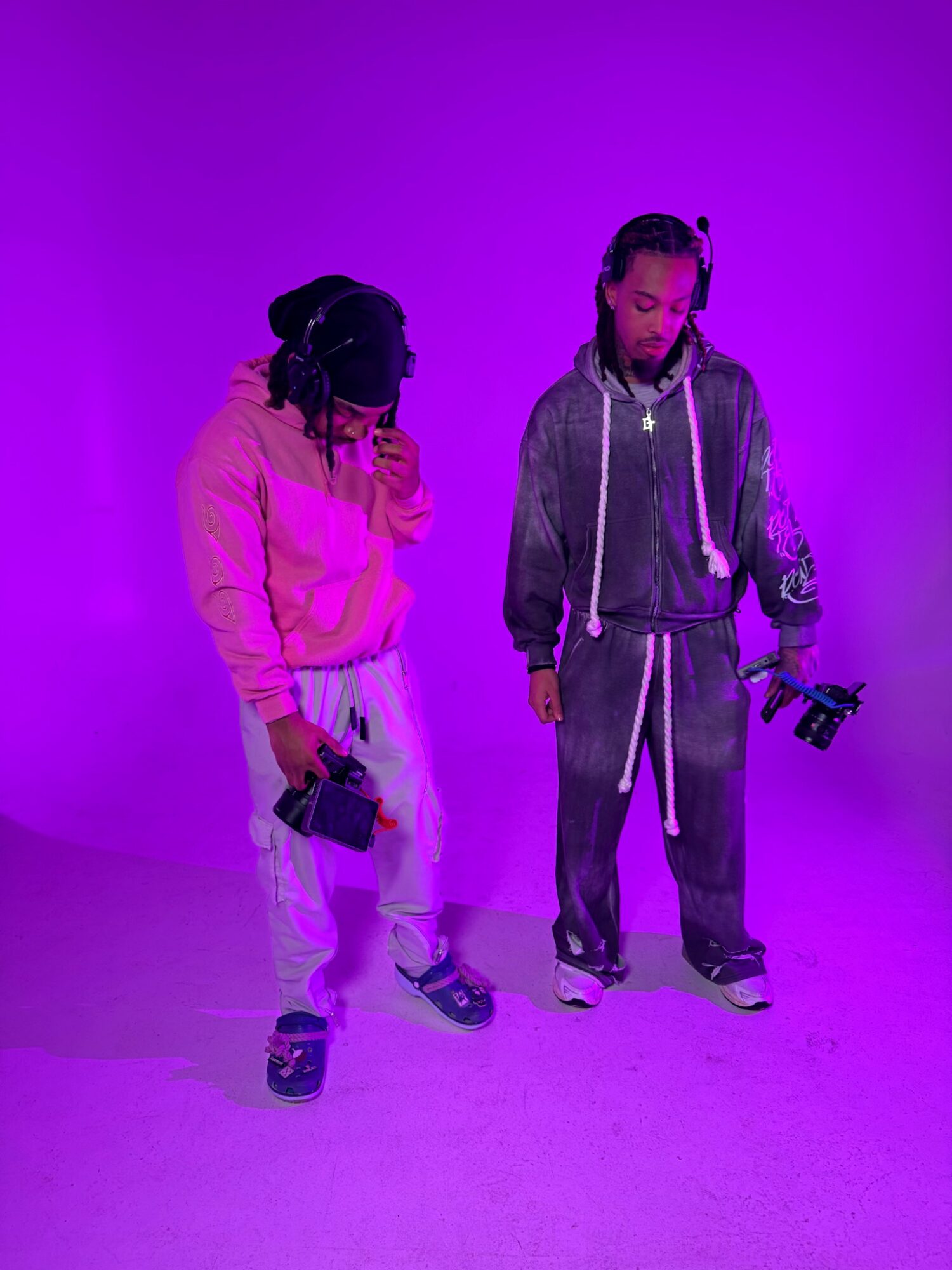 Two people stand on a purple background, each holding a camera, one wearing a pink hoodie and the other a gray tracksuit.