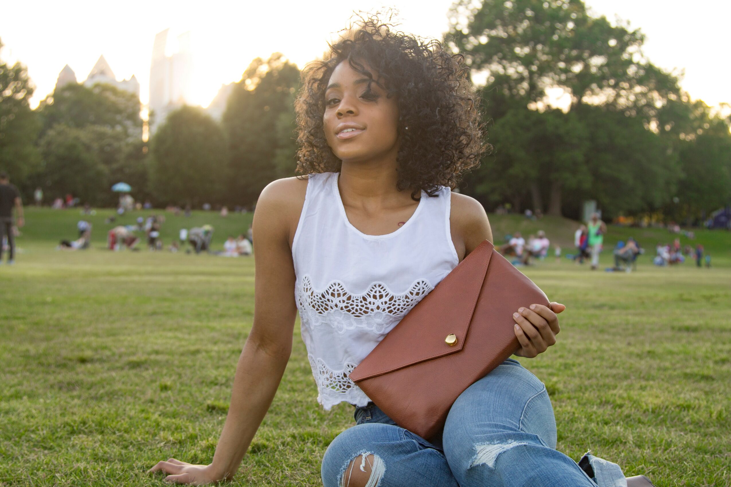 Woman with curly hair sitting on grass holding a brown clutch bag in a park with trees and people in background.