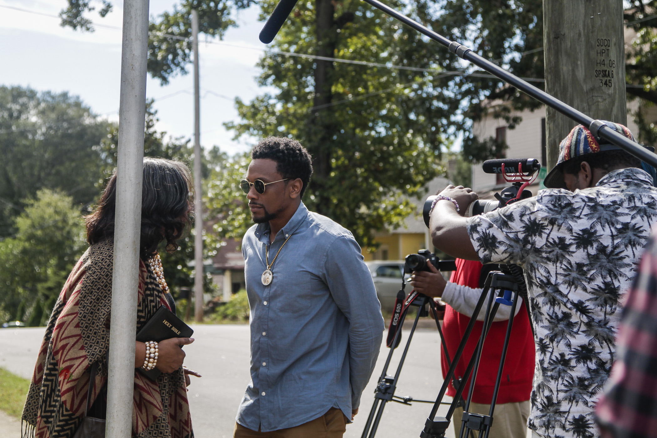 Two people engaged in conversation outdoors, with a camera crew filming them, trees and a street in the background.