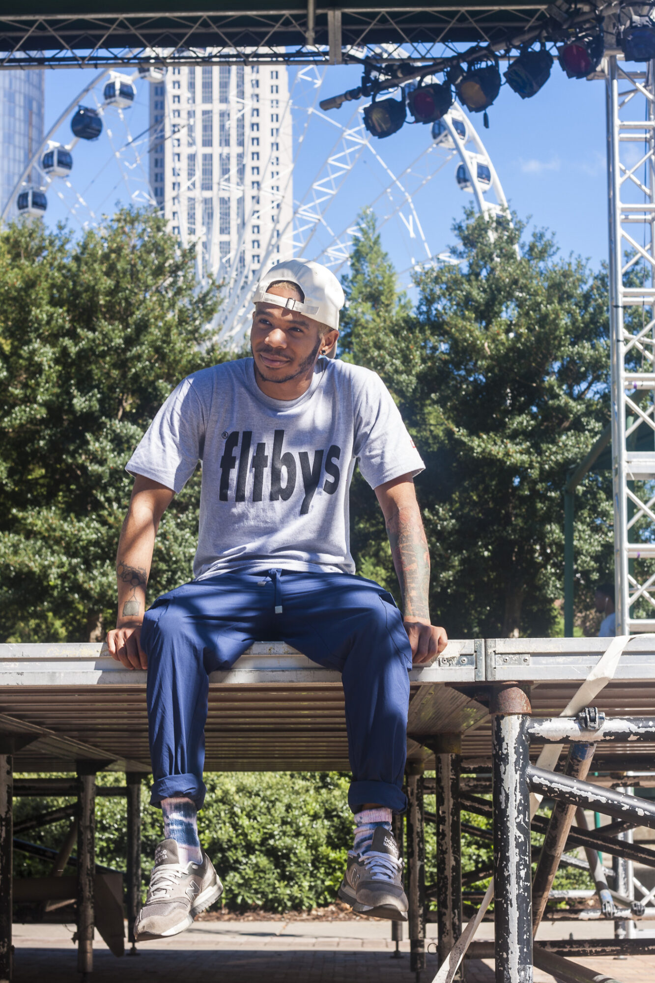 Person sitting on a stage outdoors with trees and a Ferris wheel in the background, wearing a white t-shirt and cap.