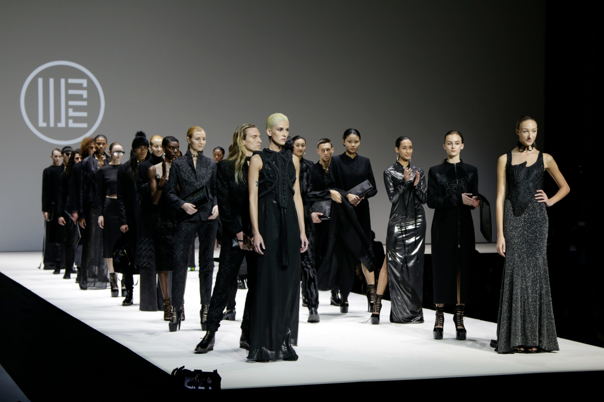 Group of models walking on runway in black and metallic outfits, with a logo projected on the wall behind them.