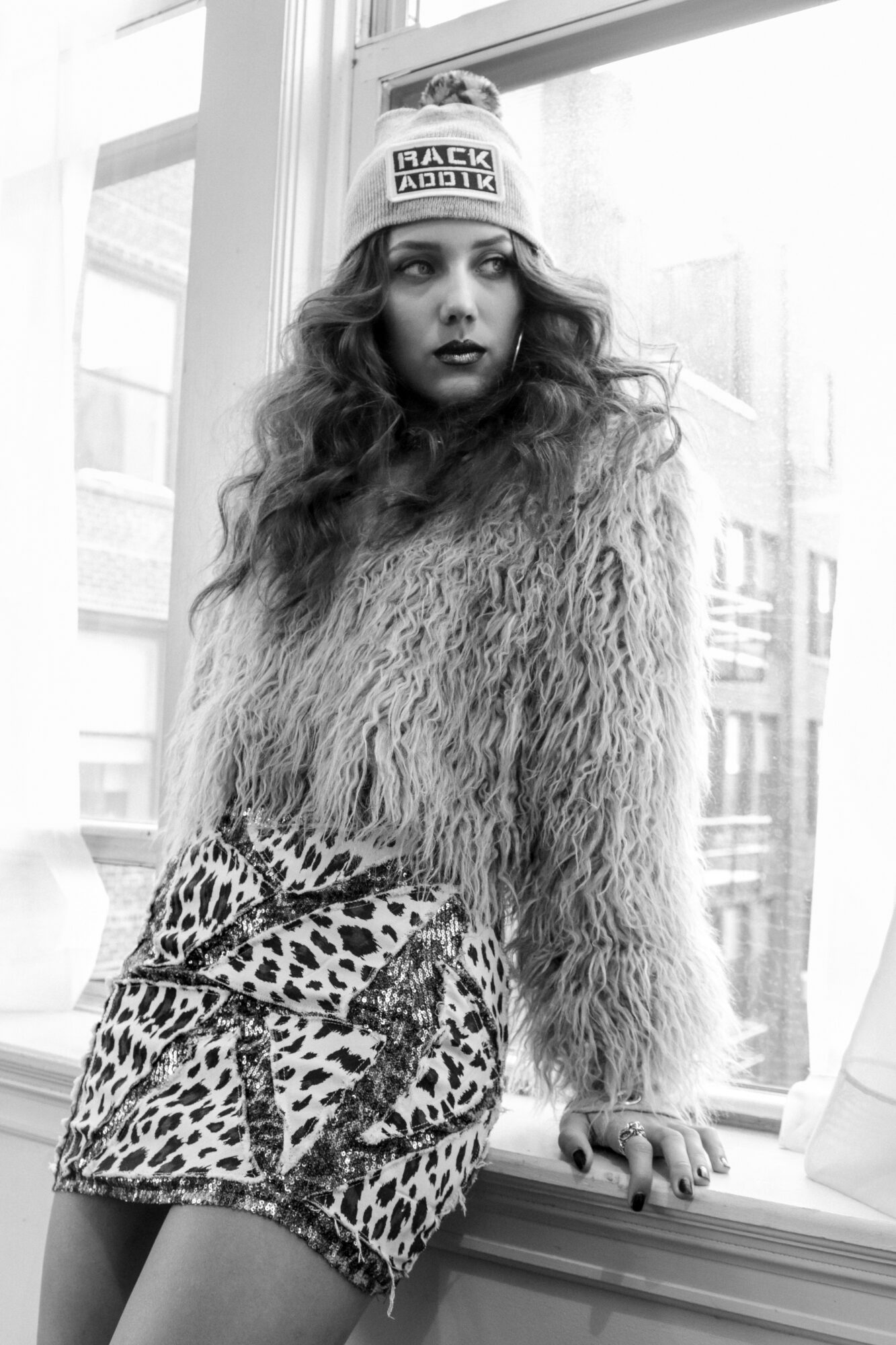 Young woman with long curly hair wearing a beanie, furry sweater, and leopard print skirt, sitting by a window.
