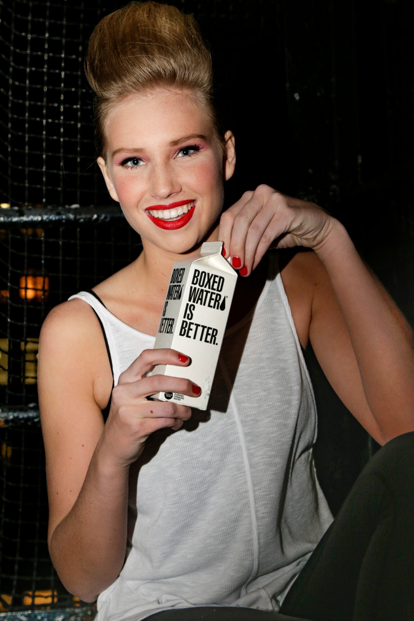 Young woman with blonde hair in a bun, smiling, holding a carton with a straw, wearing a white tank top.