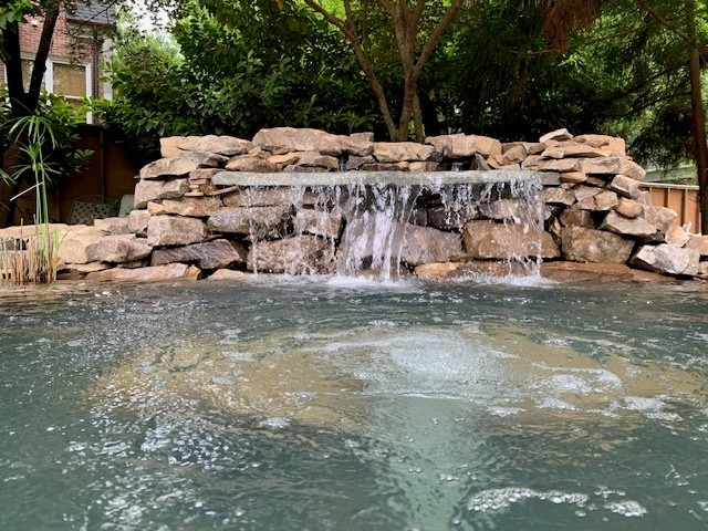 Water flows over a stone waterfall into a pool, surrounded by trees and greenery.