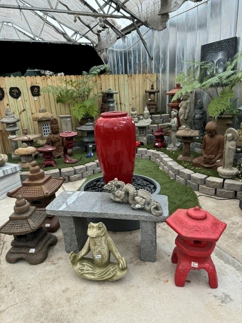 Garden with statues, plants, and decorative objects, including a large red vase and a stone table with a frog sculpture.