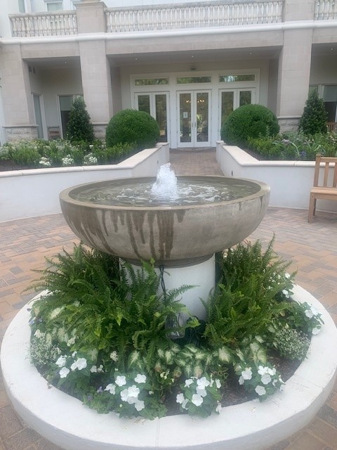 A round fountain with water spouting in front of a building entrance, surrounded by green plants and white flowers.