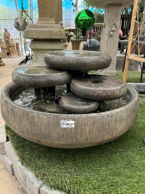 Stone fountain with multiple rounded basins stacked, water flowing, surrounded by artificial grass, outdoor setting with plants and structures.
