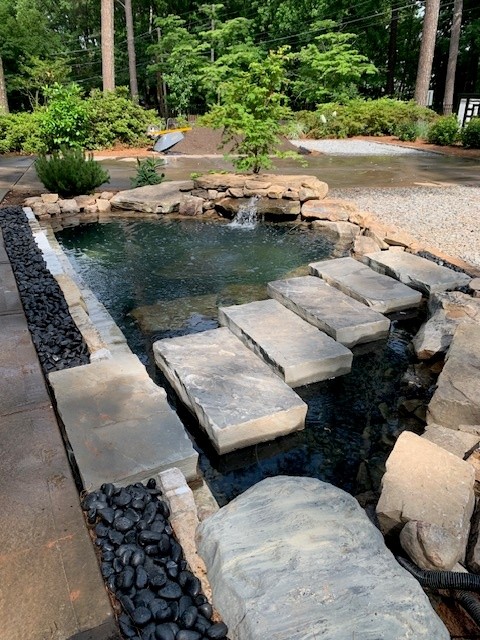 Stone pathway crosses a small pond with rocks and water plants, surrounded by trees and garden landscape.