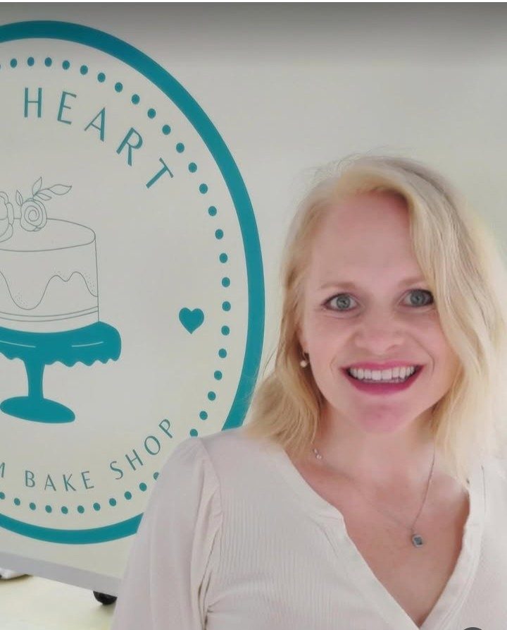 Smiling woman with blonde hair standing in front of a sign with a cake illustration and the words 'HEART BAKE SHOP'.