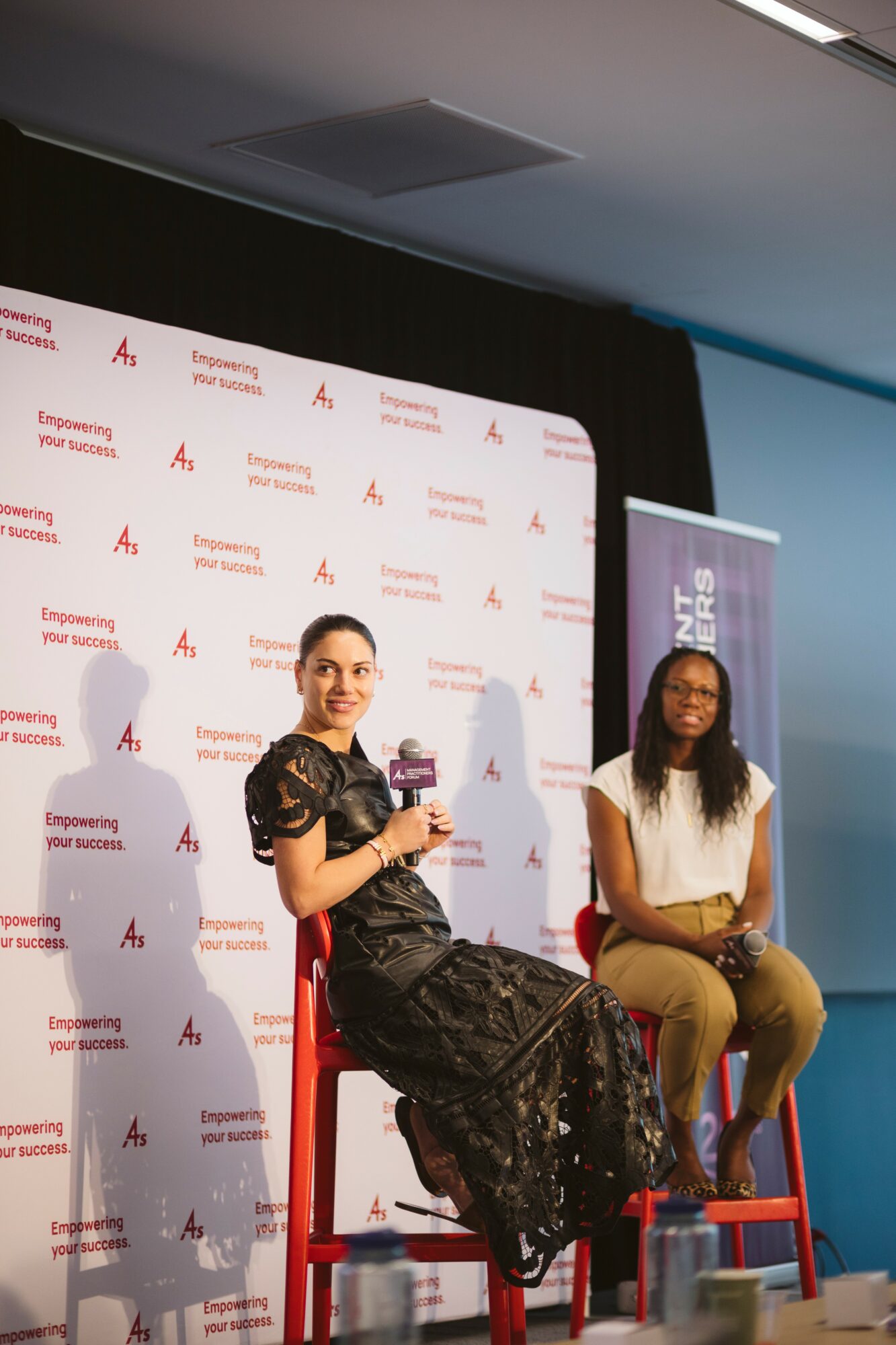 Two women sit on red chairs on a stage with a backdrop and a banner, one holding a microphone, smiling.