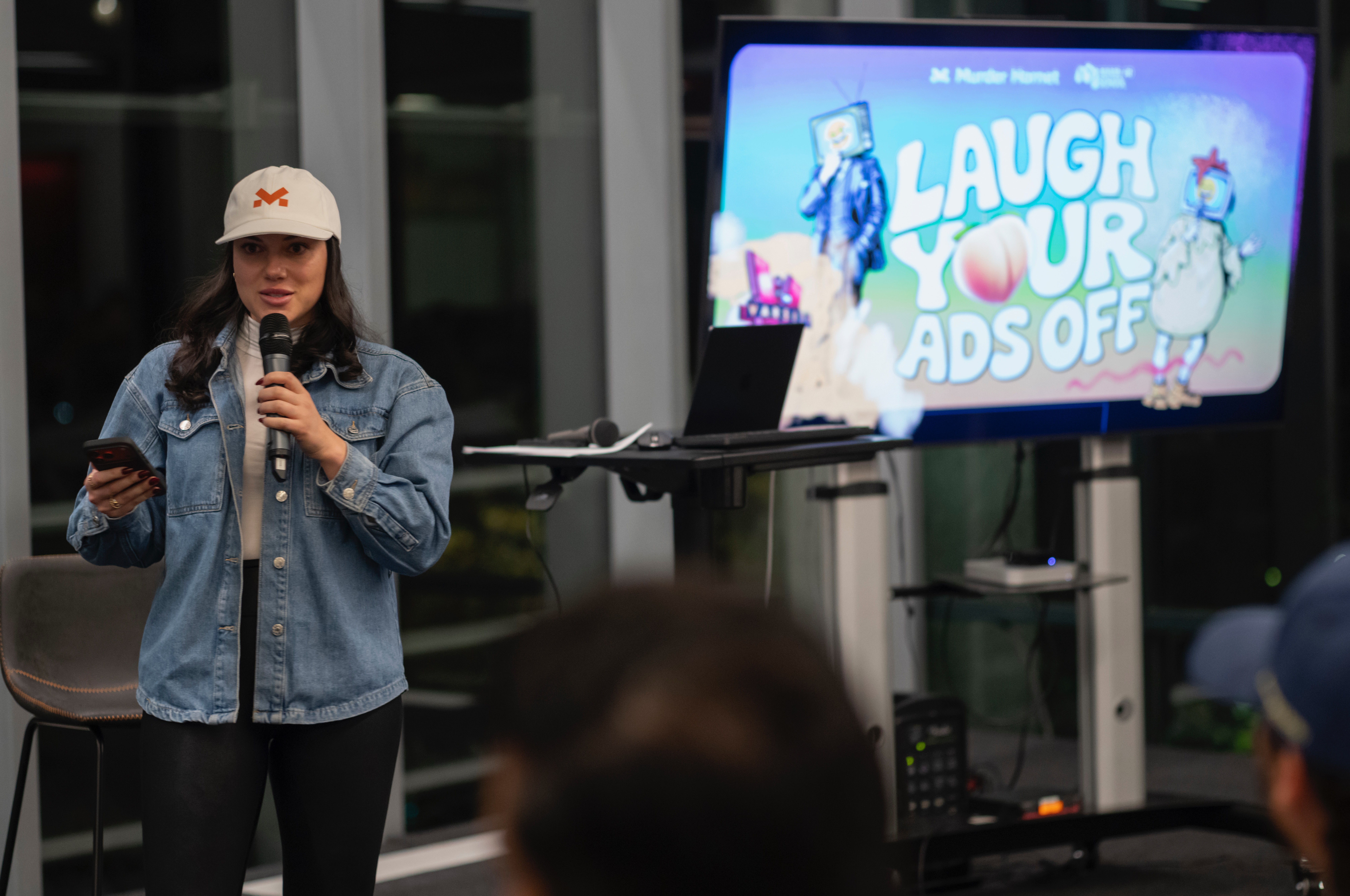 Young girl wearing a white cap and denim jacket speaking into a microphone, with a screen displaying 'Laugh Your Ads Off' behind her.