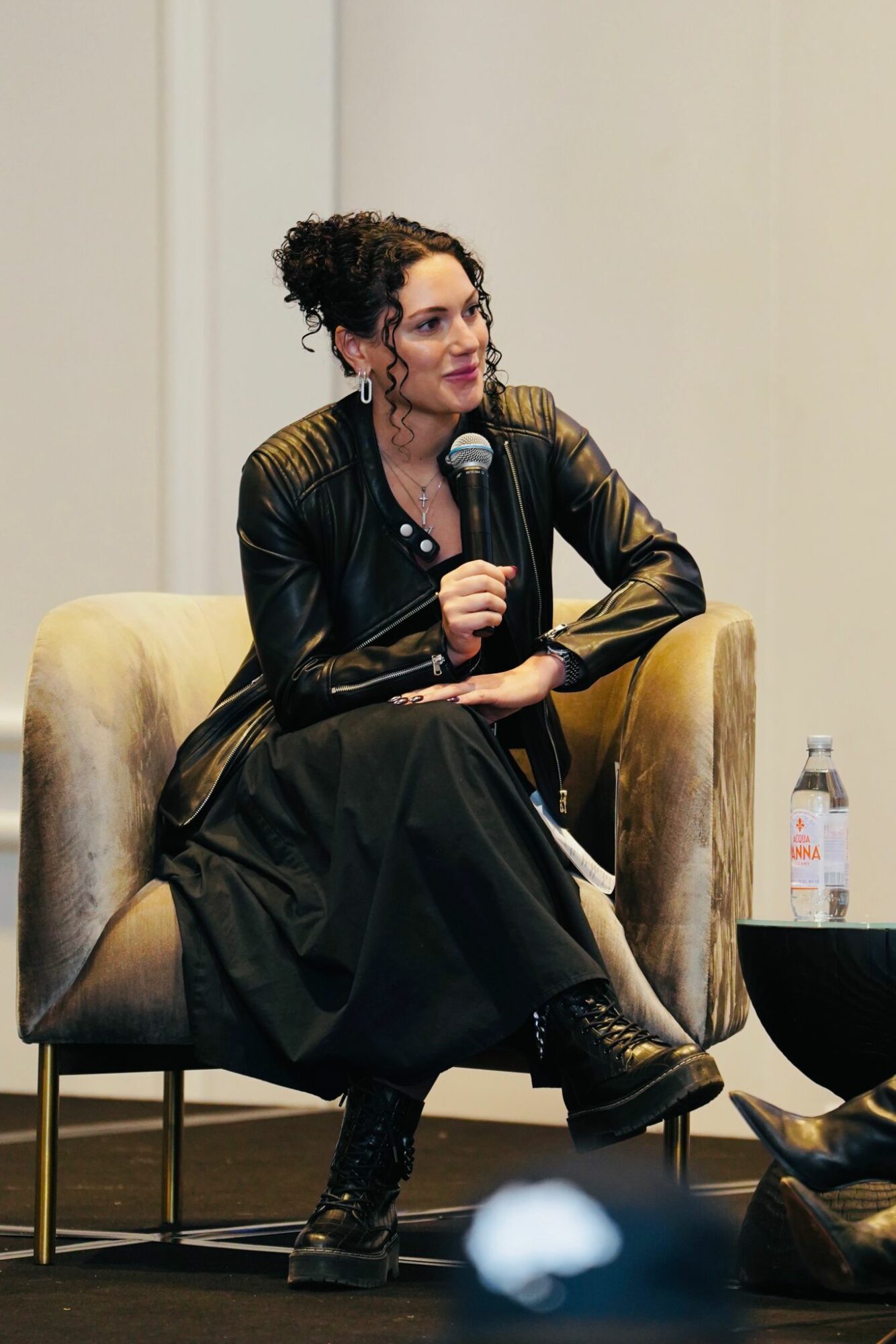 Woman with curly hair in a black leather jacket and dress, sitting on a beige armchair, holding a microphone, with a water bottle nearby.