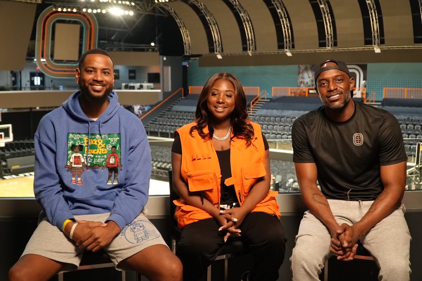 Three people sitting in an indoor arena, smiling, with empty seats and a stage in the background.