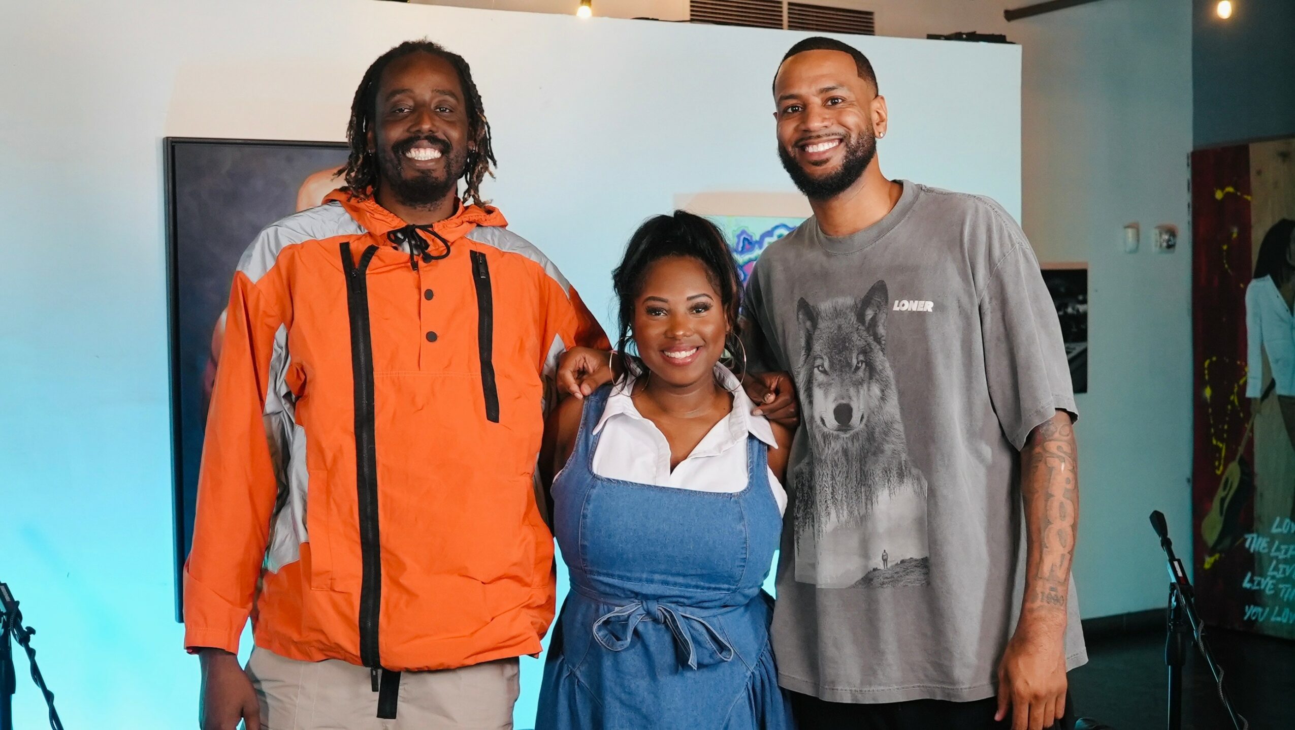 Three people smiling indoors, standing close together, with a blue background and artwork behind them.