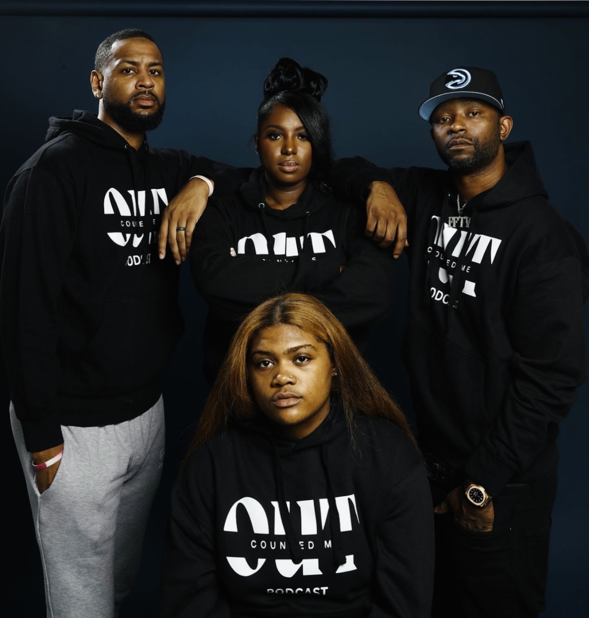 Four people wearing black hoodies with white text, standing together indoors against a dark background.