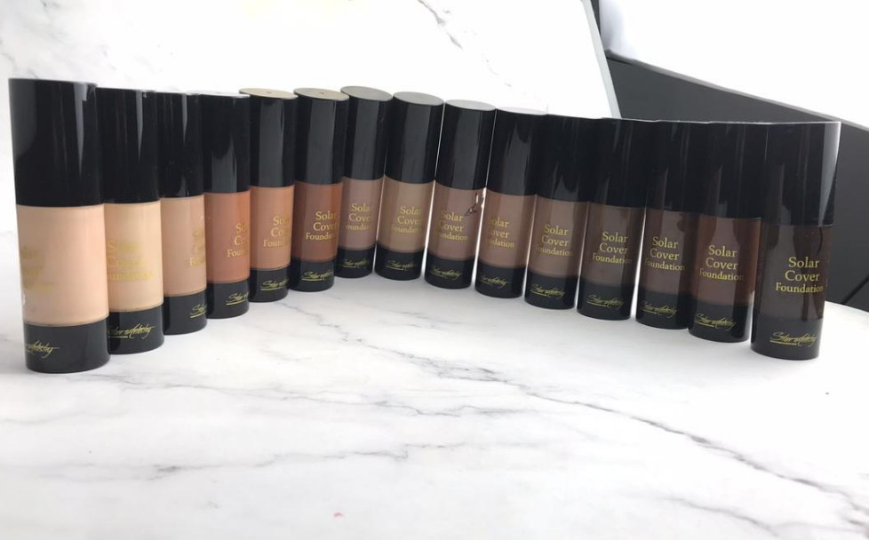 Row of 16 skincare or cosmetic bottles with black caps, beige labels, and gold text, arranged on a white surface.