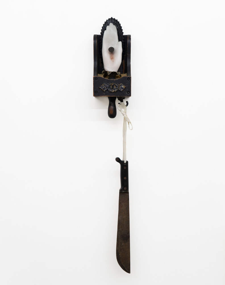 Wall-mounted object resembling a vintage clock with a knife hanging below, against a plain white background.