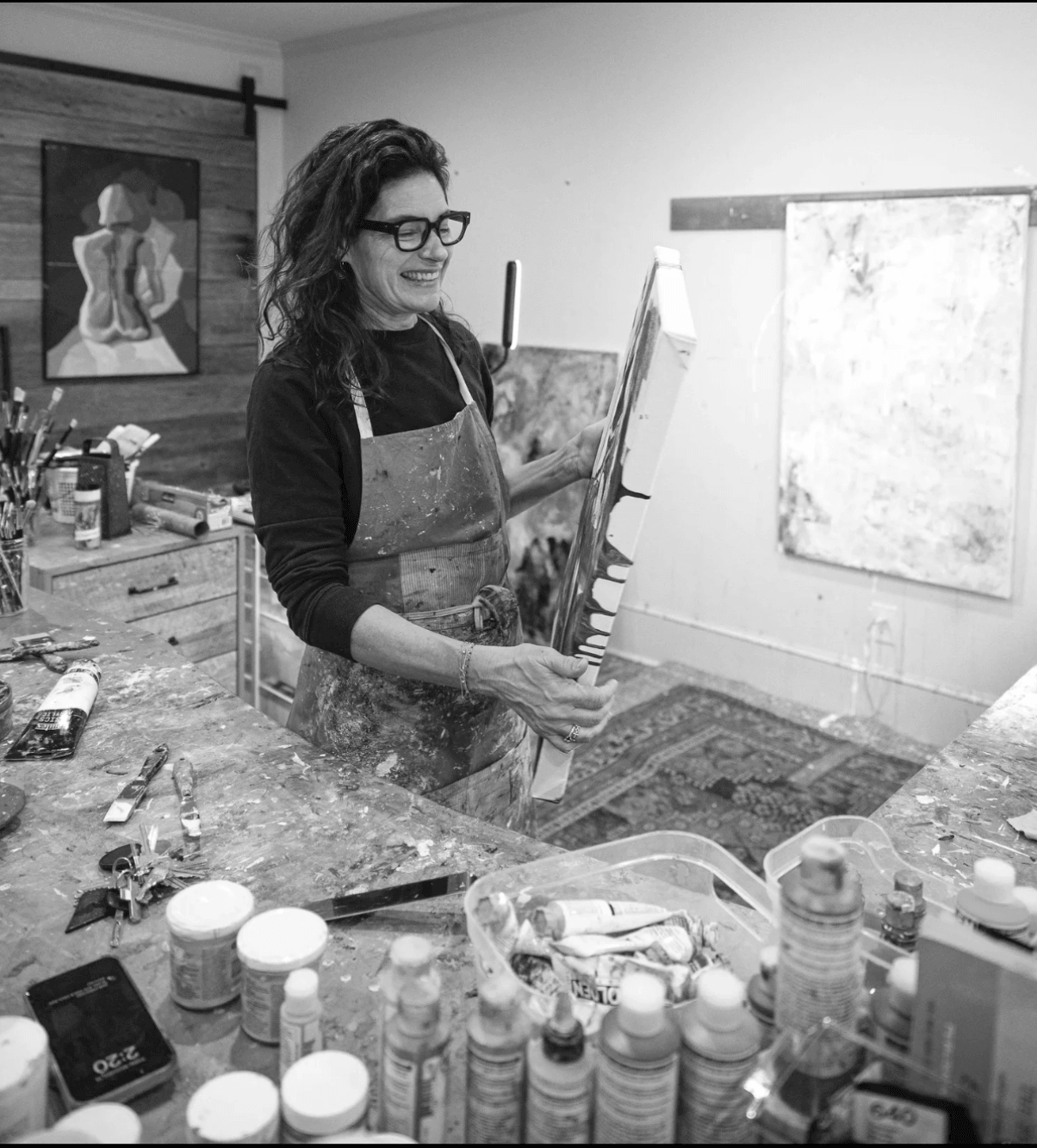 Woman with glasses and apron painting in art studio, surrounded by art supplies and paintings.