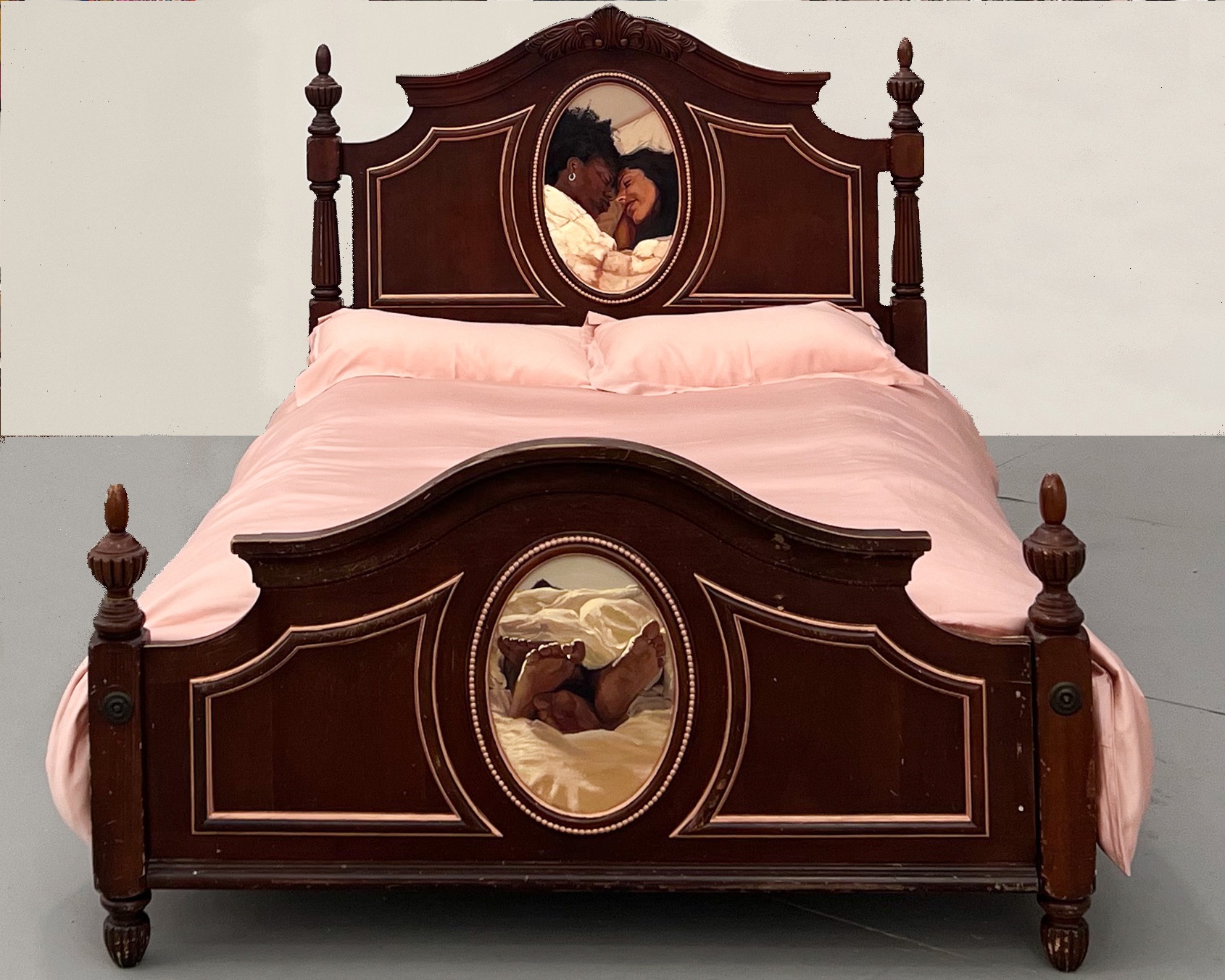 A wooden bed with a headboard and footboard, featuring oval portraits of a couple, pink bedding, and a plain background.