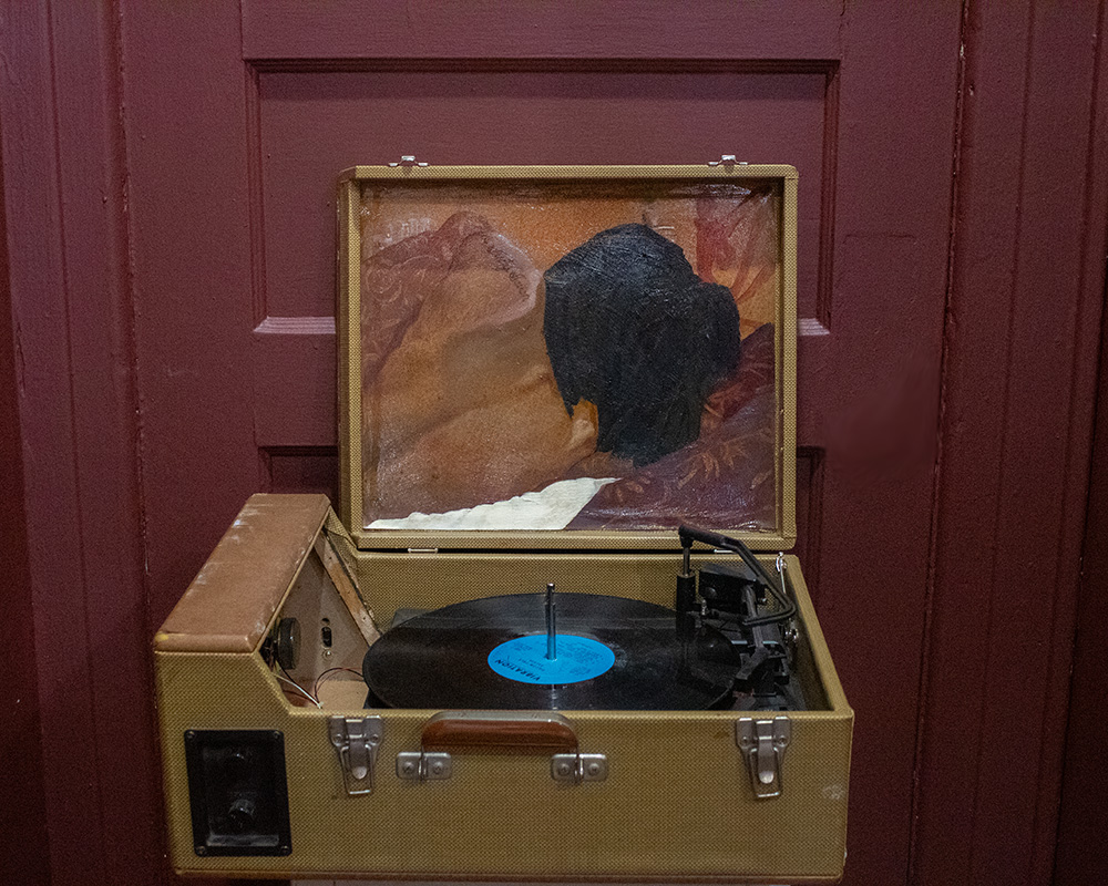 A vintage record player with a painting of a person's head on the lid, set against a red wooden background.