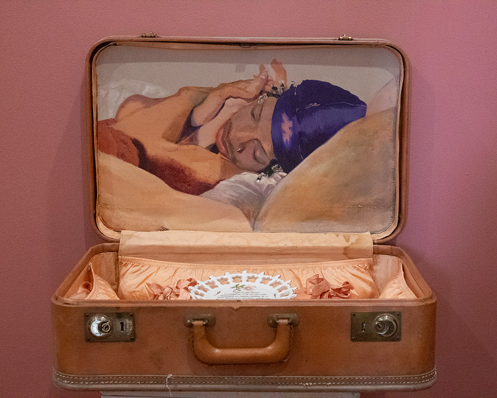 Open suitcase with a painting of a person sleeping, resting on a pillow, inside. Pink background. Alt text is 20 words.