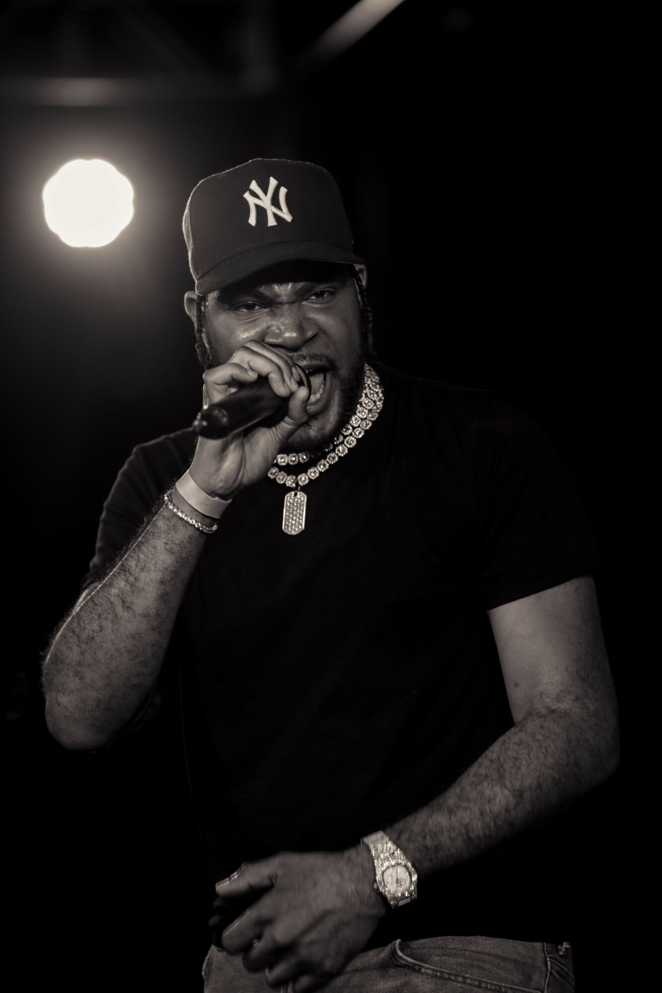 Person wearing a New York Yankees cap, holding a microphone, with jewelry, on stage in black and white.