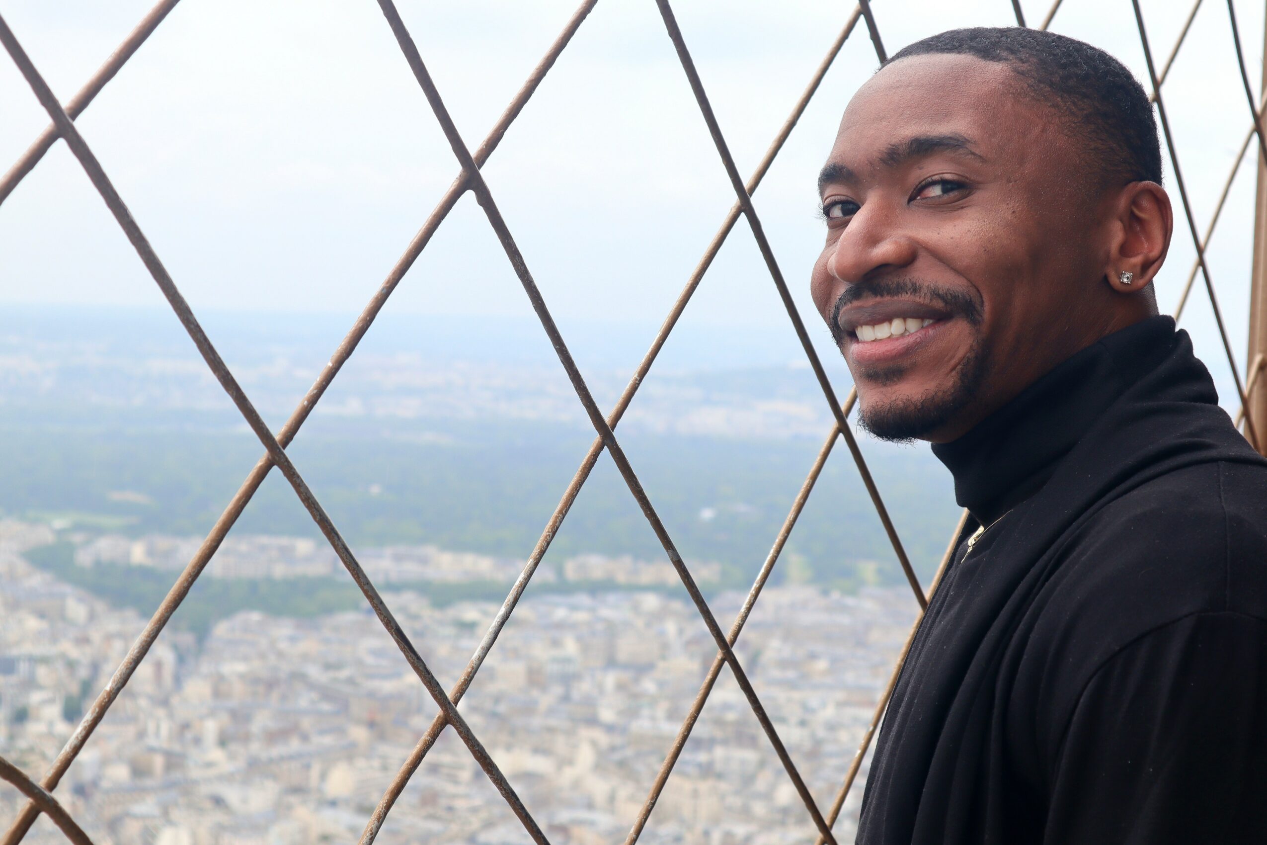 Smiling man in black jacket on a high observation tower with a cityscape background.