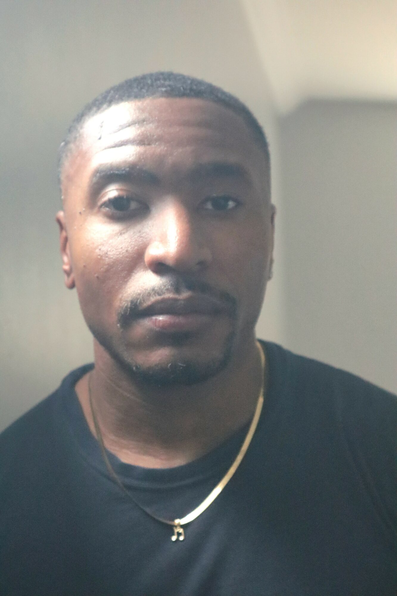 Portrait of a man with short hair, wearing a black shirt and a gold chain, looking at the camera.