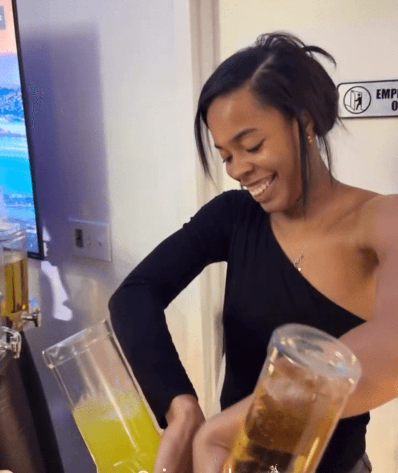 Smiling woman with short dark hair in a black top pouring a yellow drink into a glass, in a room with a sign on the wall.