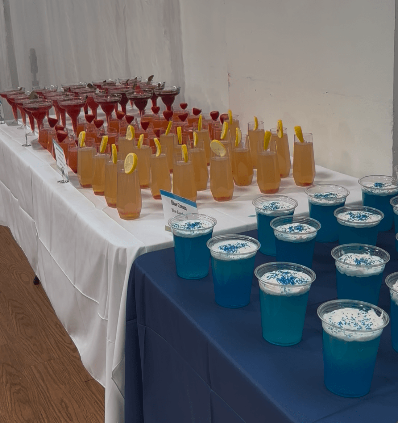 Table with colorful drinks in cups and glasses, arranged in rows, with a white and blue tablecloth, against a plain wall.