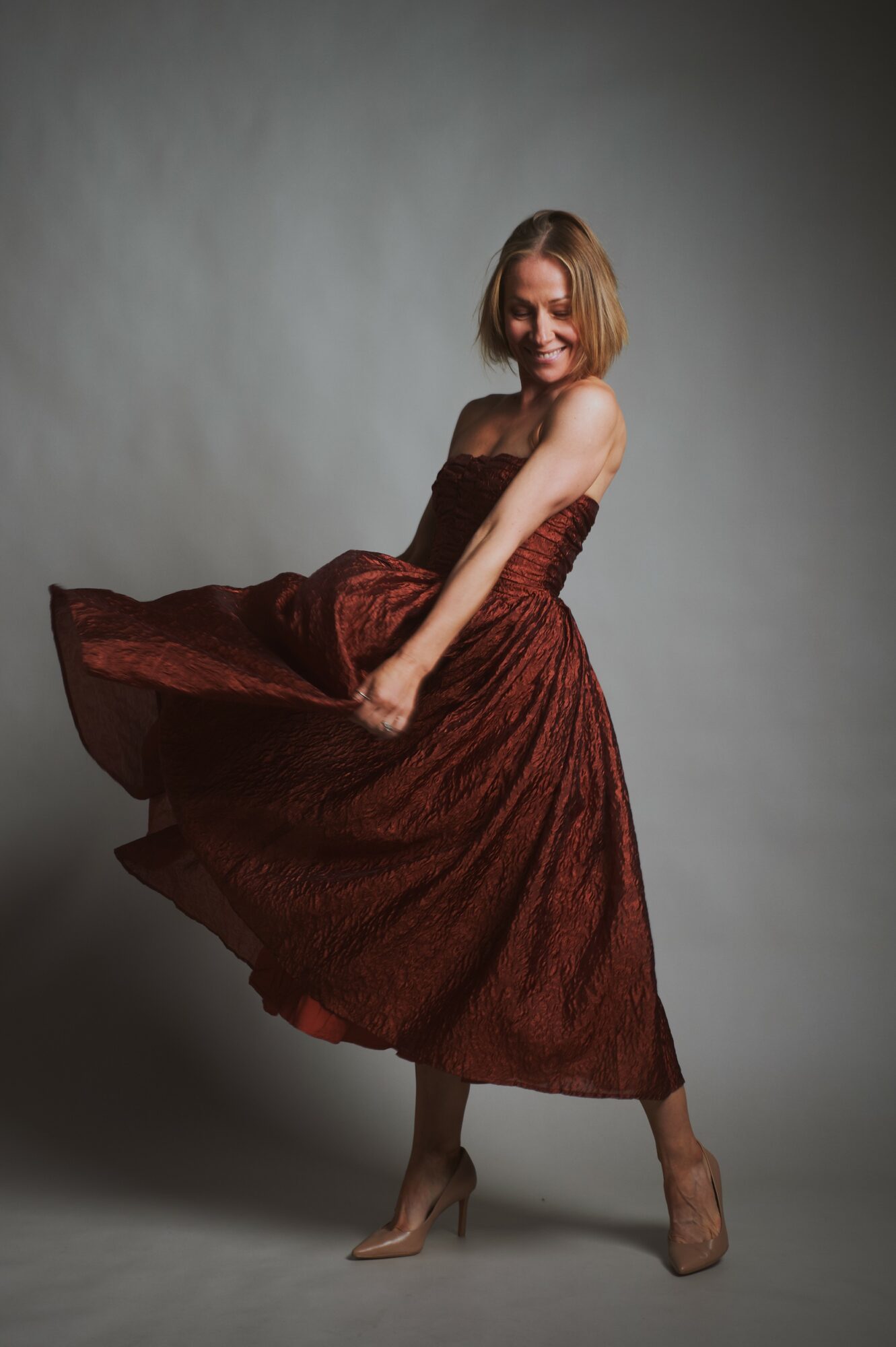 Woman in a flowing, strapless, brown dress, smiling, holding the skirt, standing on a gray background.