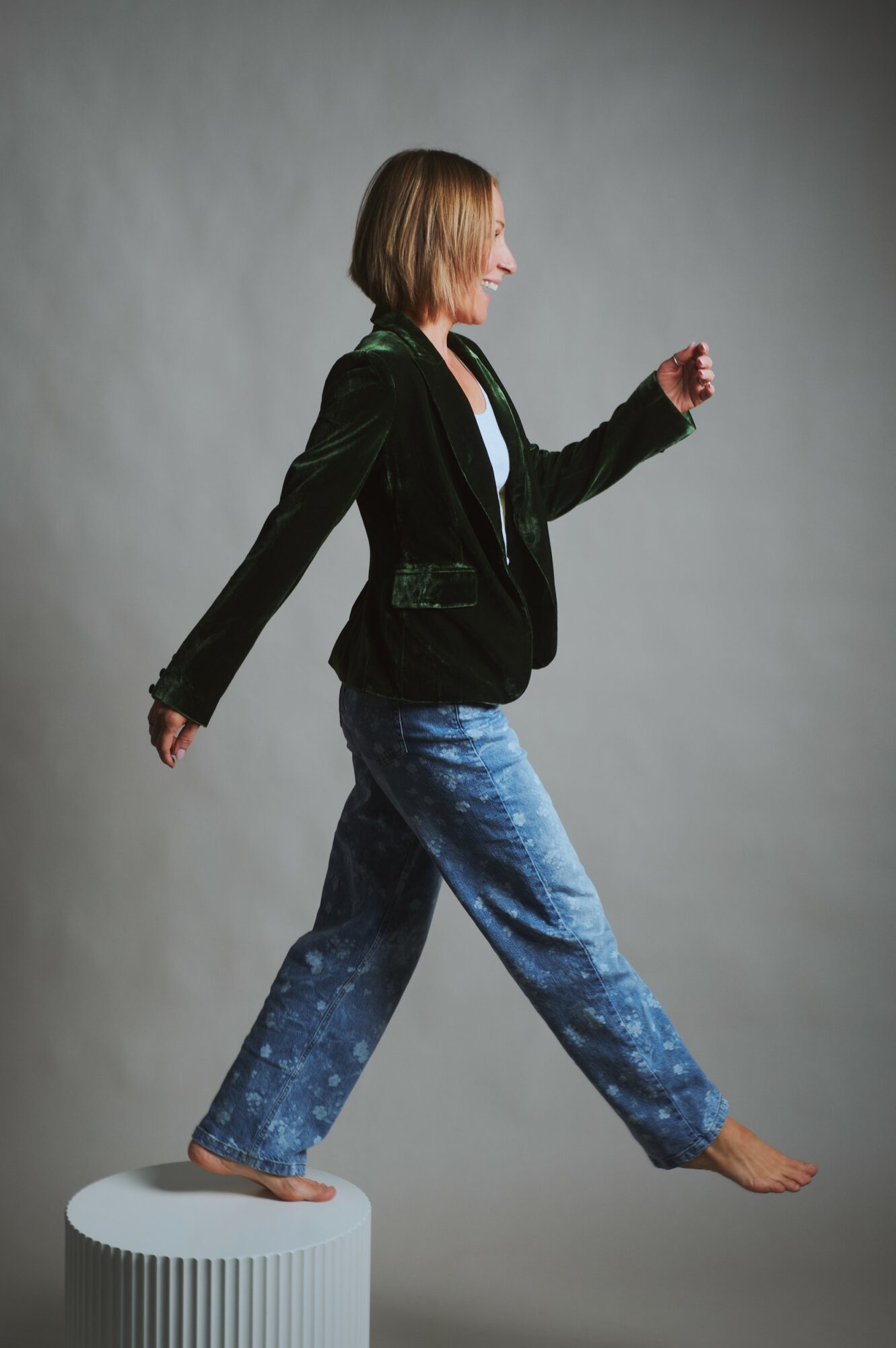 Woman walking on a cylindrical platform, wearing a black jacket and blue jeans, against a plain gray background.