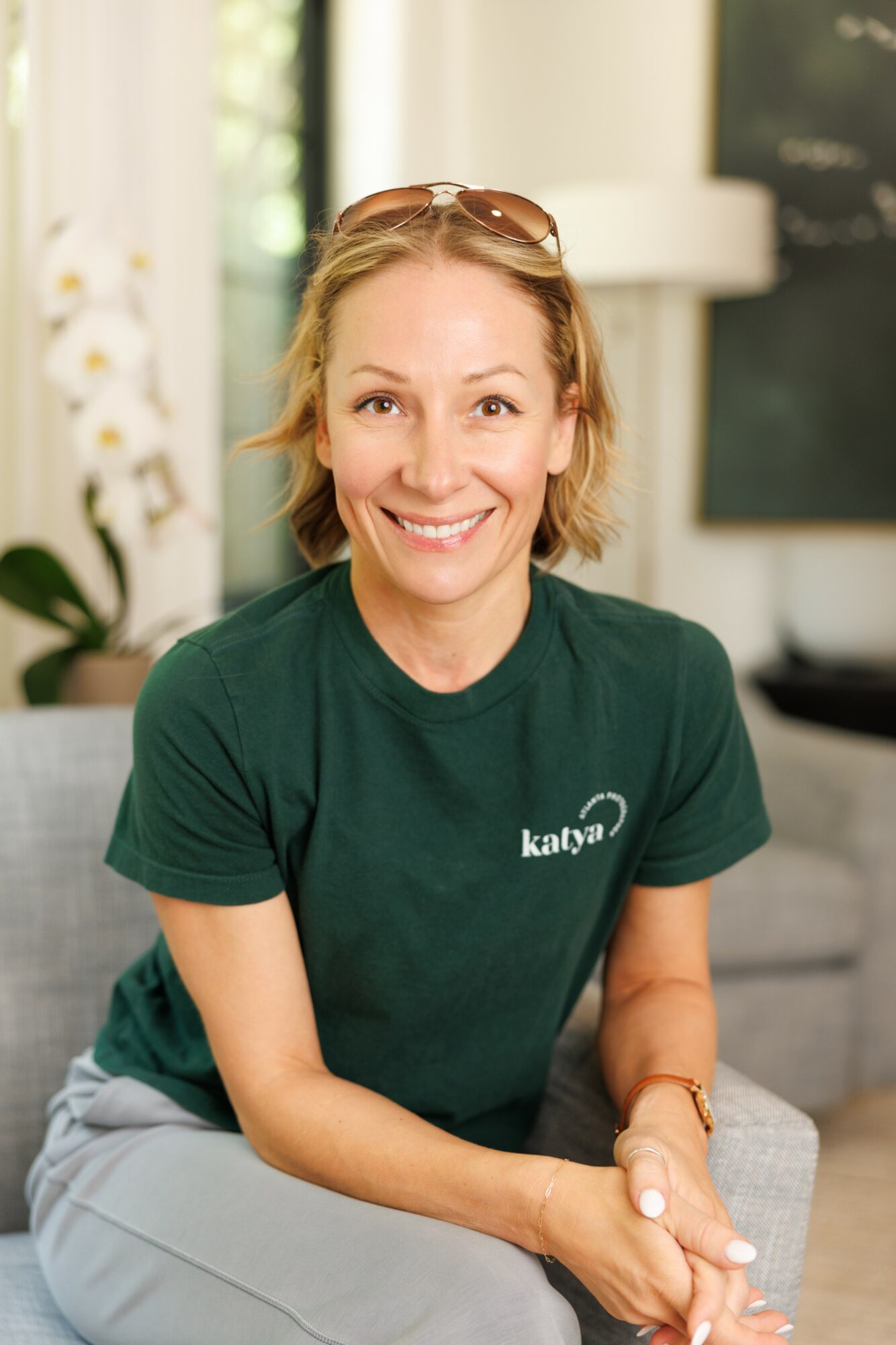 Smiling woman with short blonde hair wearing a green shirt with a logo, sitting indoors with a blurred background.