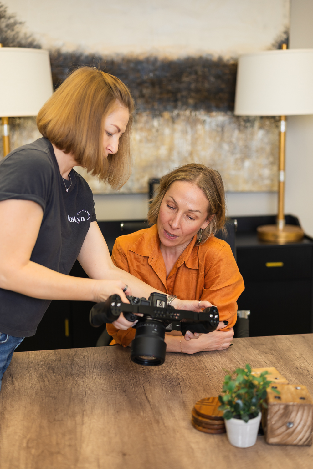 Two women look at a camera on a table, one standing and one sitting, in a room with lamps and artwork.