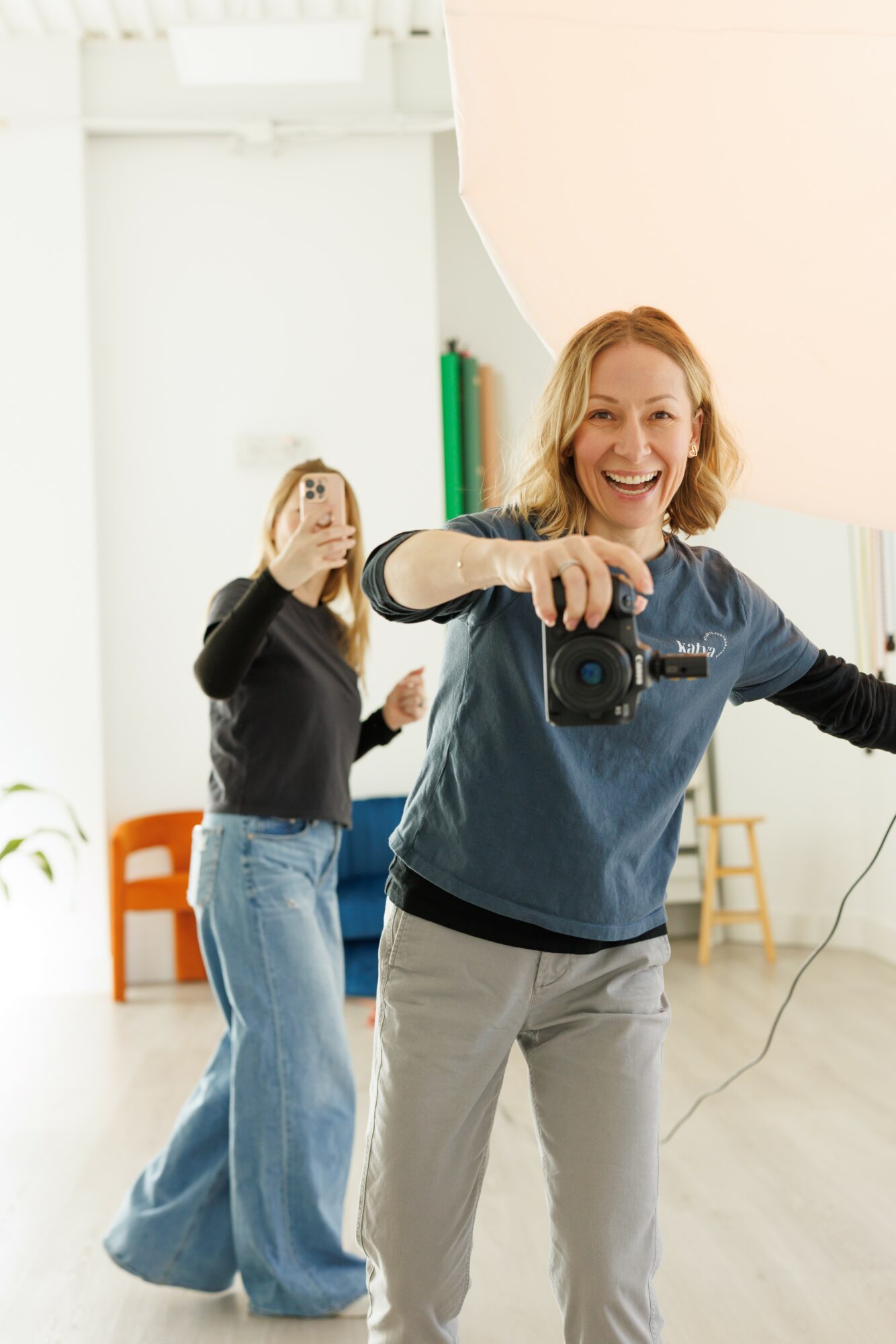 Two women smiling, one taking a selfie with a camera, in a bright room with white walls and colorful furniture.