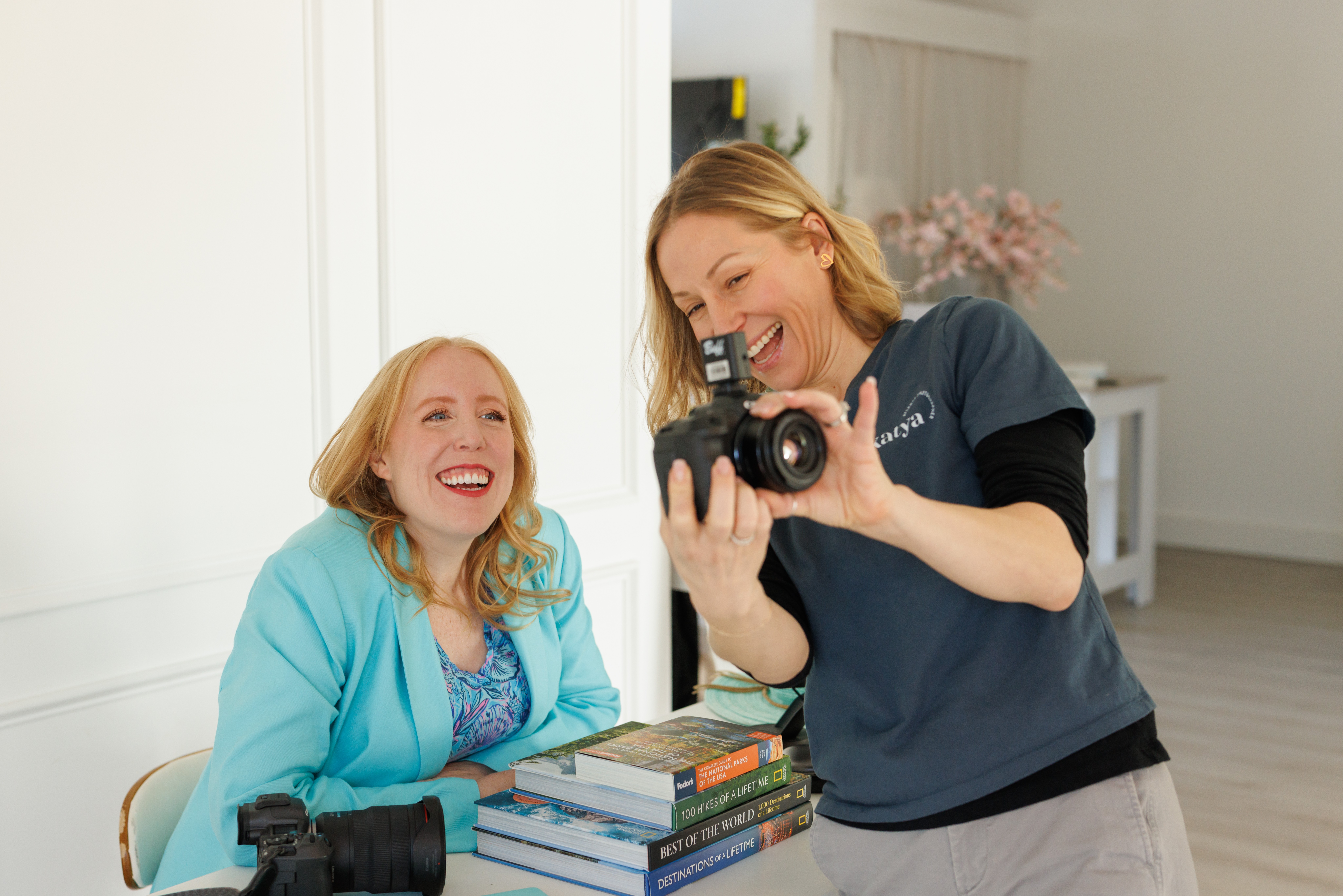 Two women smiling, one holding a camera, with books on a table, in a bright room with white walls.