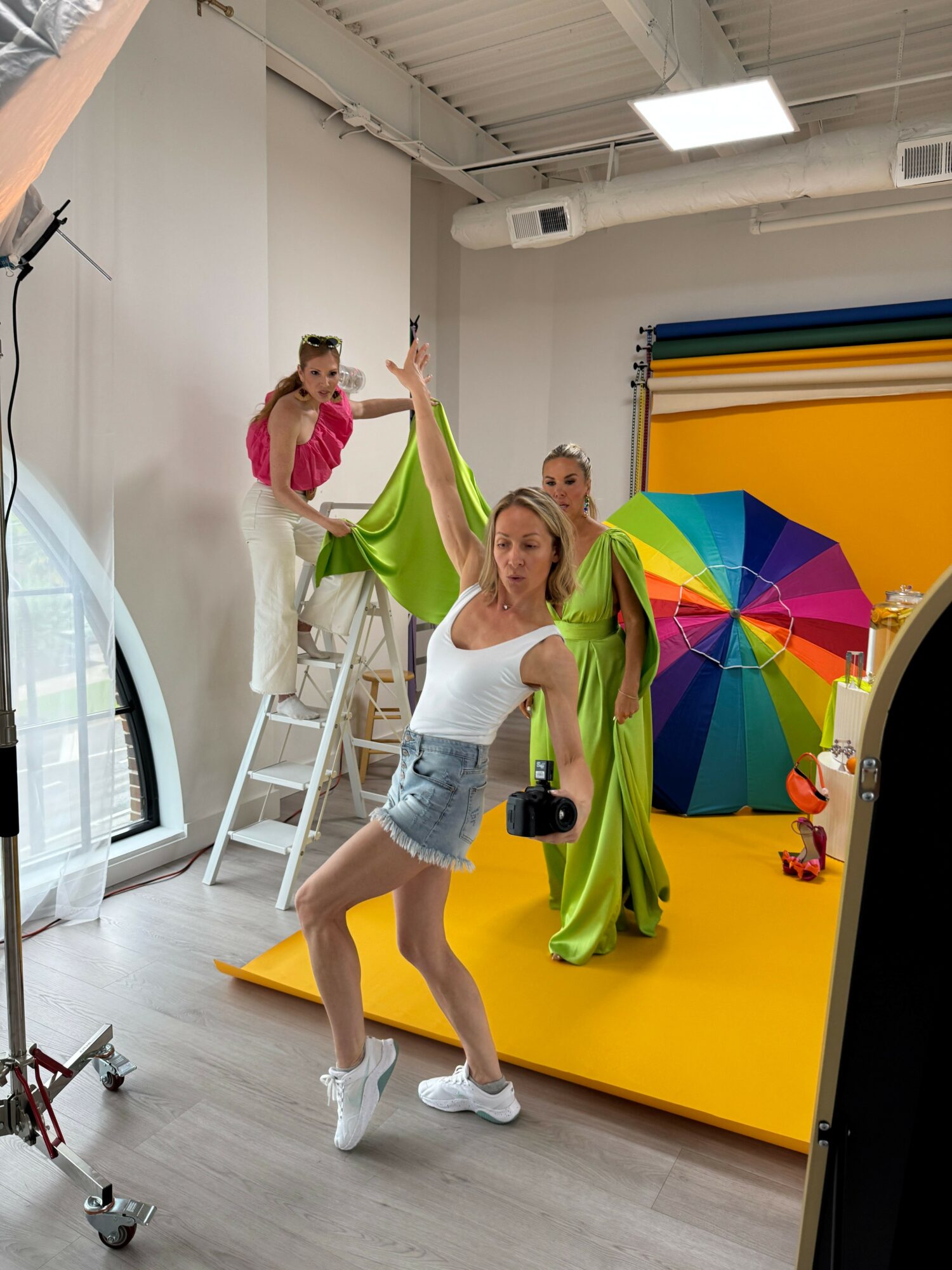 Person posing with colorful umbrella in a room with yellow wall, white ladder, and large window, holding a drink, smiling.