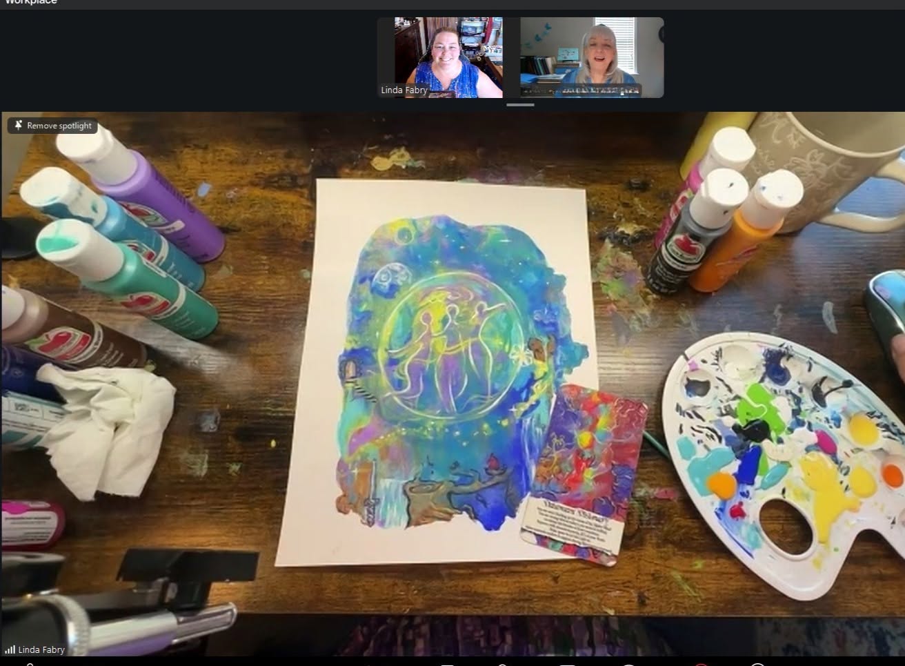 Painted artwork on paper with colorful abstract design, surrounded by paint bottles, palette, and art supplies on a wooden table.