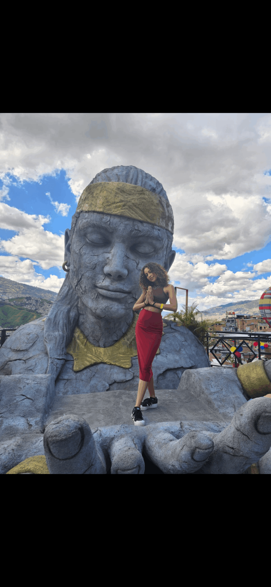 Person in red dress standing next to large gray statue of a face with a hat, outdoors with cloudy sky.