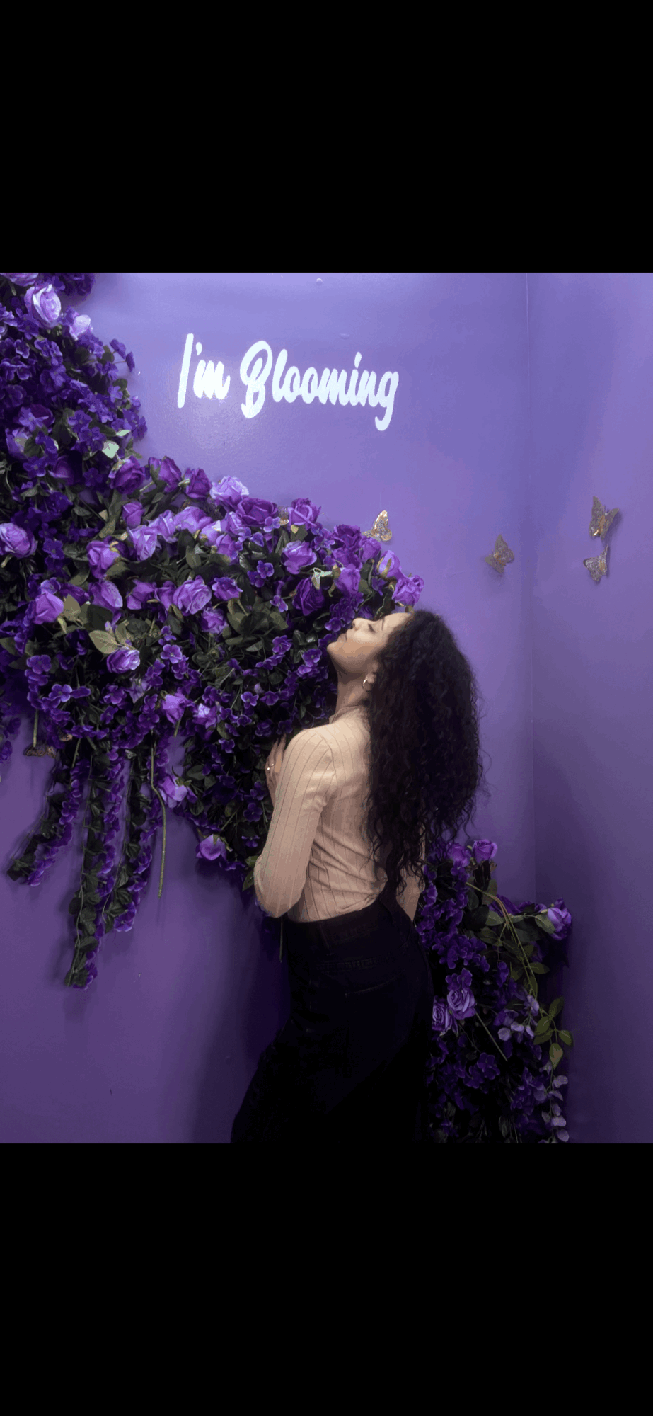 Person with long hair facing purple flower wall with neon sign reading 'I'm Blooming'.