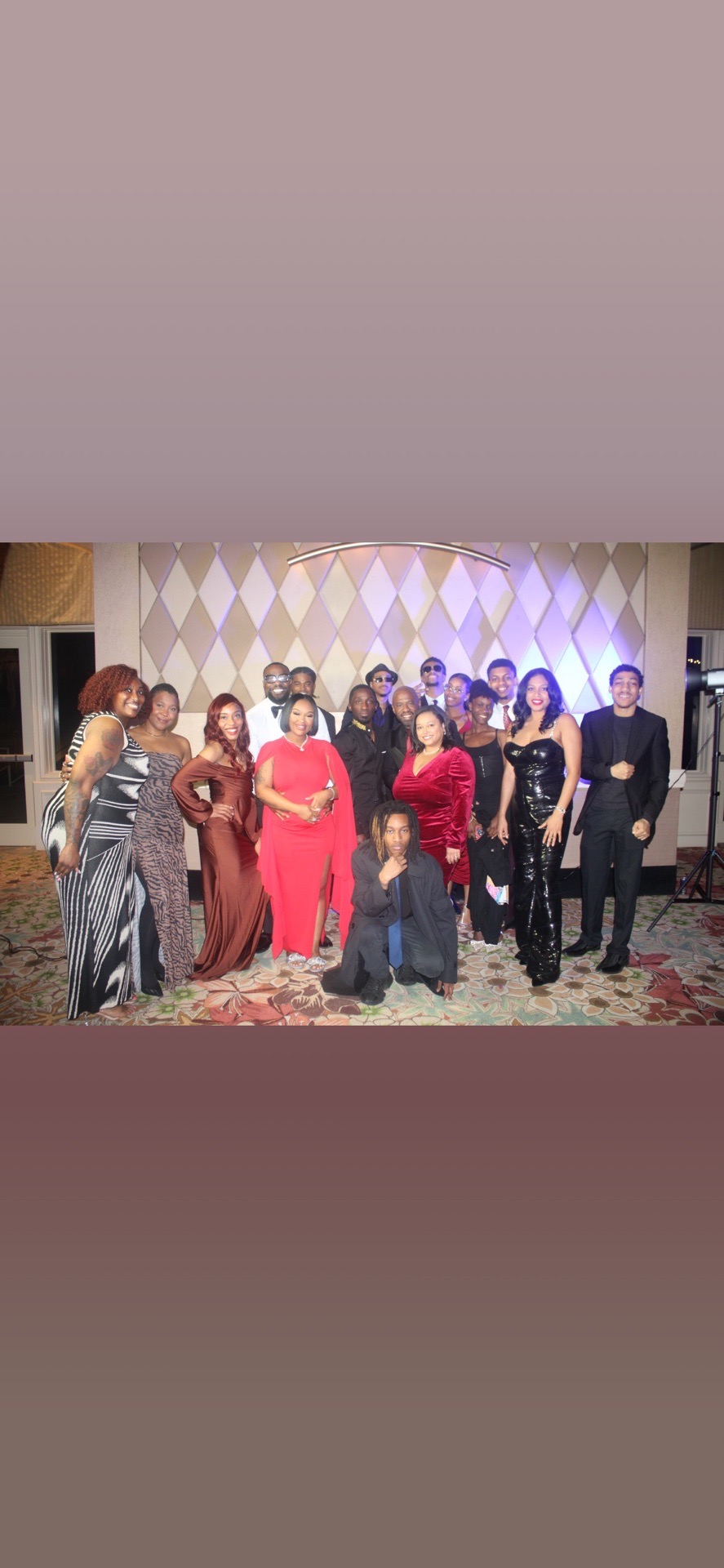Group of people posing on a stage with a patterned backdrop, some standing and some kneeling, in formal attire.