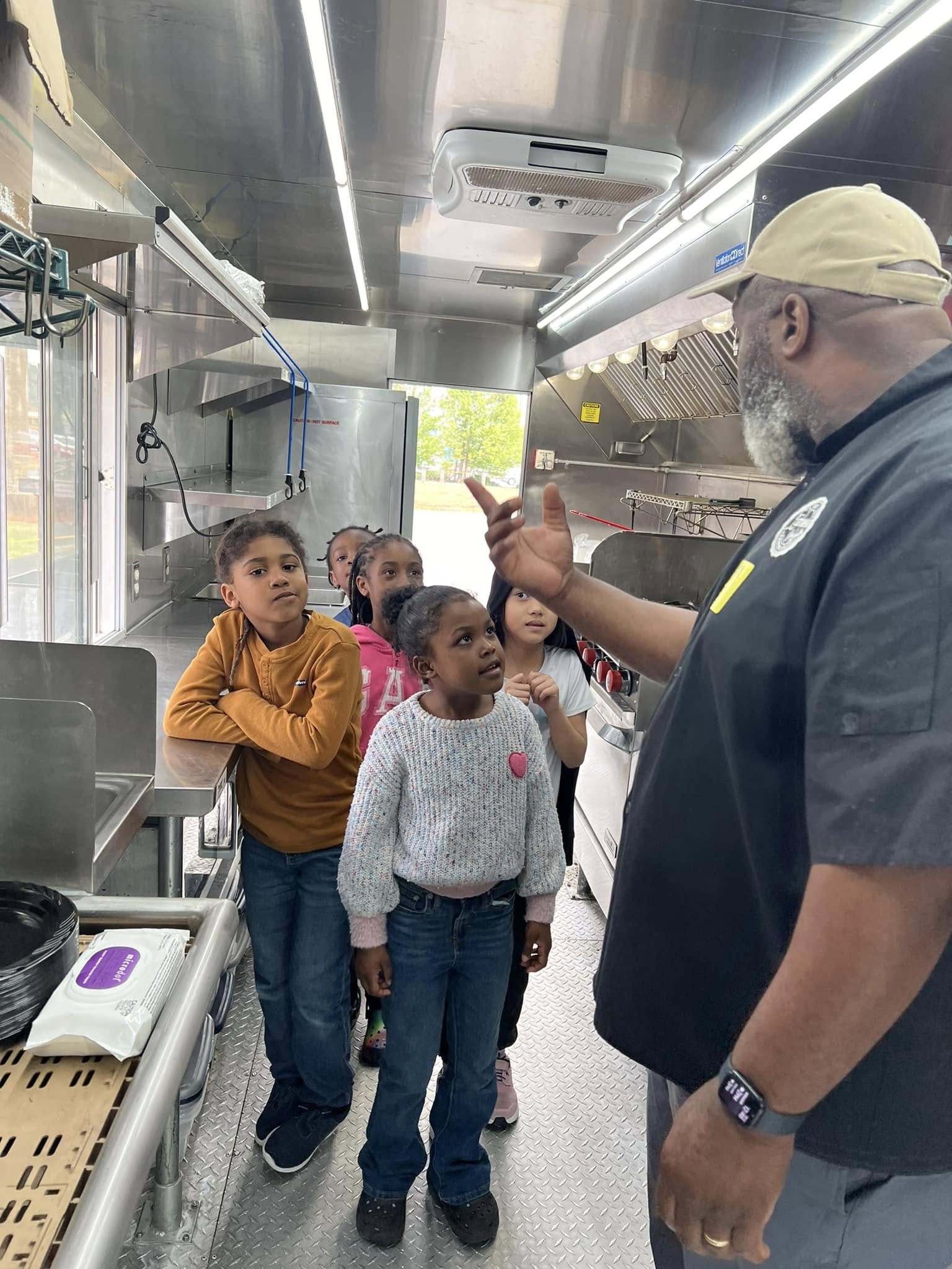 A man in uniform speaking to a group of children inside a commercial kitchen, with stainless steel appliances and bright lighting.