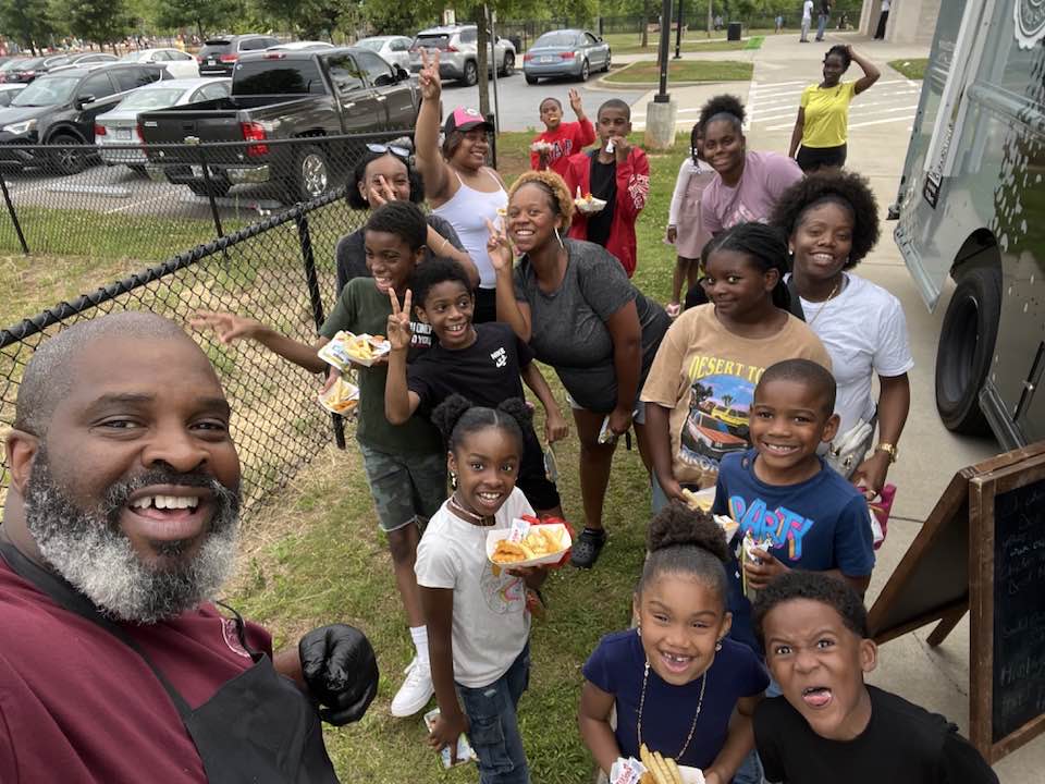 Group of children and adults outdoors near a bus, smiling and holding food, with cars and trees in background.