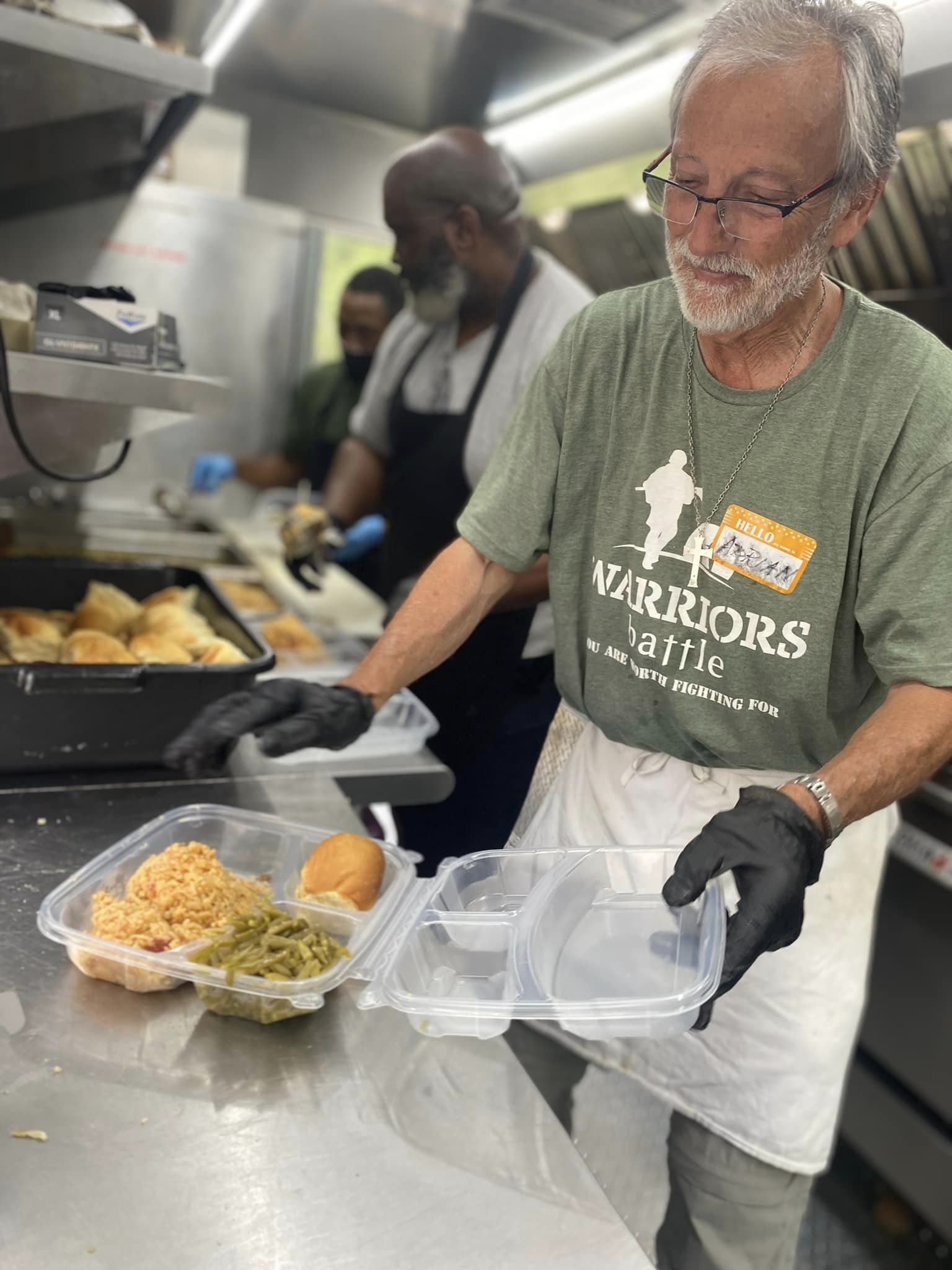 Man in green shirt preparing food in a kitchen, with two other people in the background, food containers on the counter.